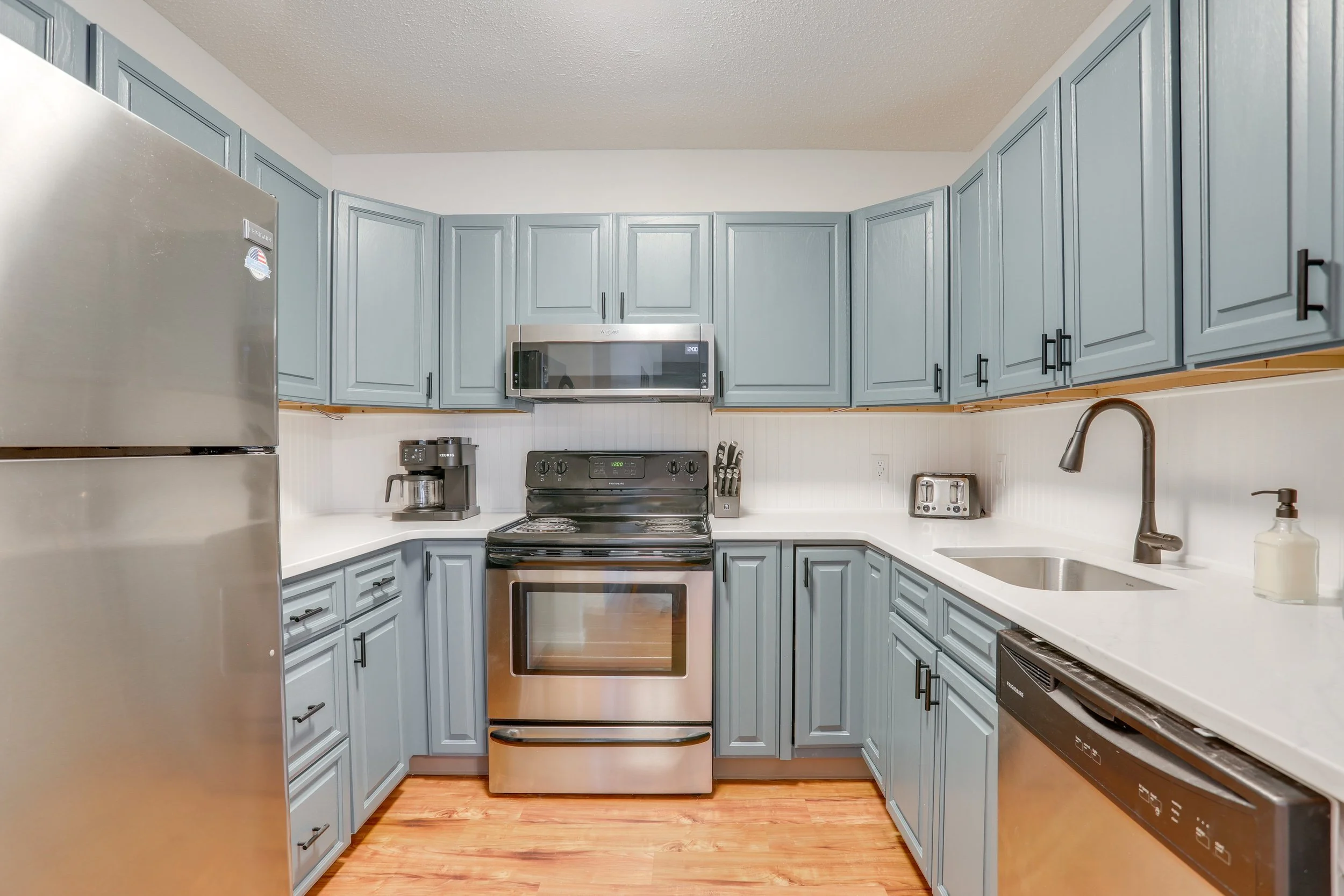 A modern kitchen with blue cabinets, a stainless steel refrigerator, black stove, microwave, toaster, coffee maker, and sink with a dark faucet, on a white countertop.