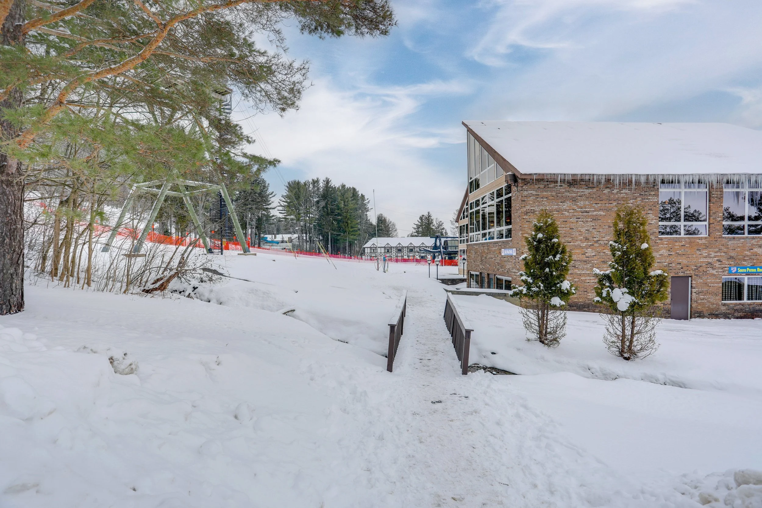 A snow-covered outdoor scene with a brick building, small evergreen trees, a pathway, and a blue sky with clouds.