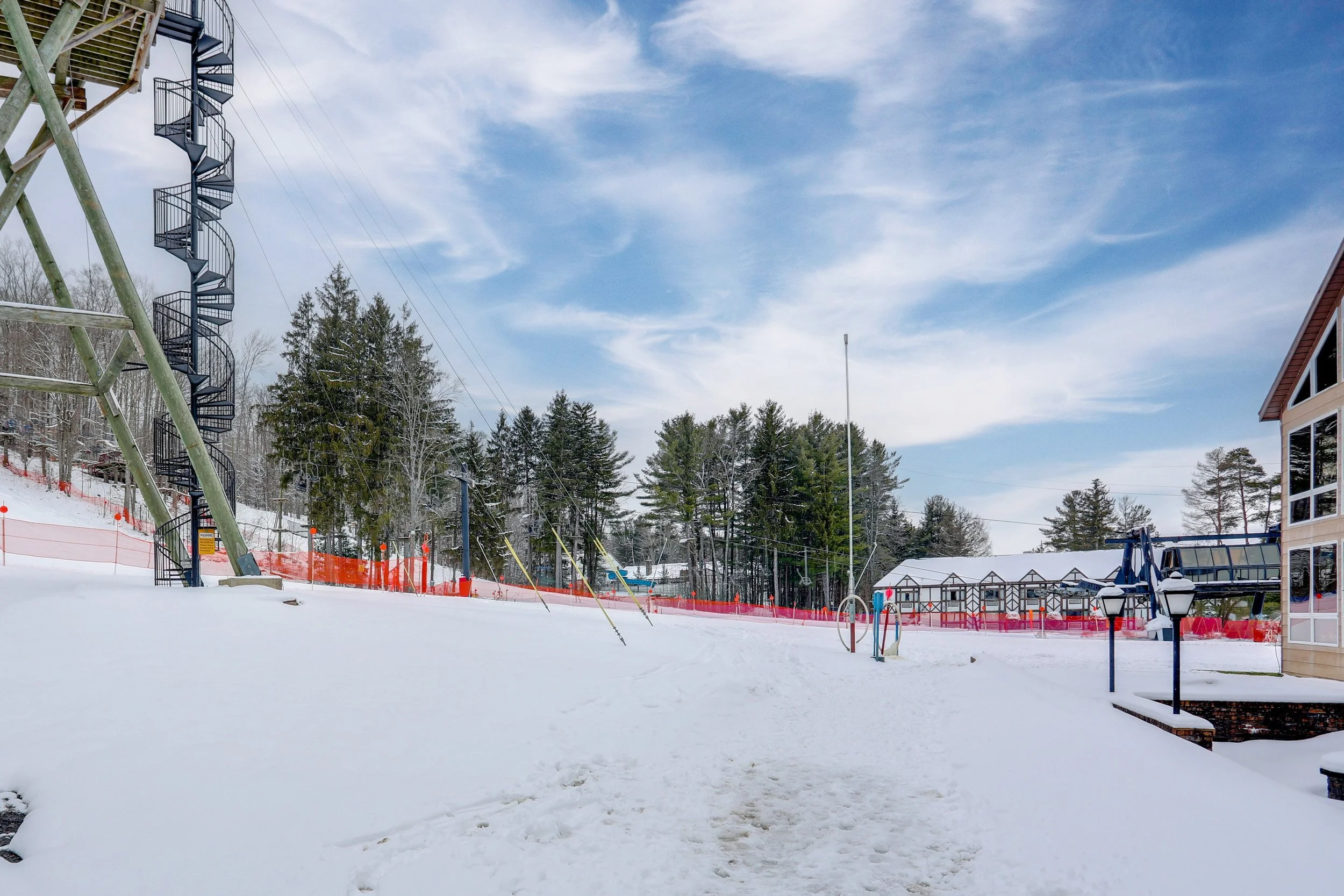 Snow-covered ski resort with ski lift tower on the left, ski trails marked with orange fencing, and trees in the background under a partly cloudy sky.