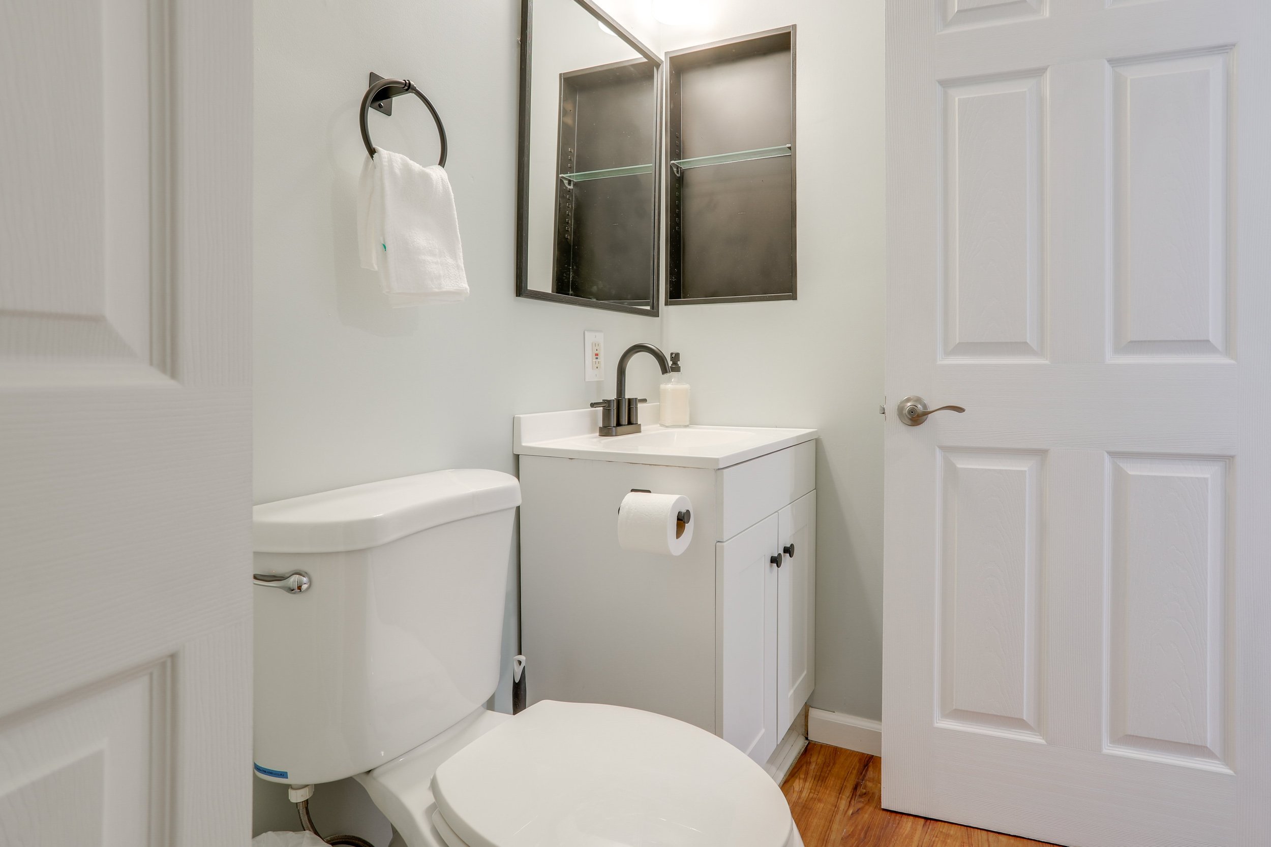 Small bathroom with white walls, a white toilet, a white cabinet with a sink, a black-framed mirror, a black towel ring with a white towel, a black medicine cabinet, and a wooden floor.
