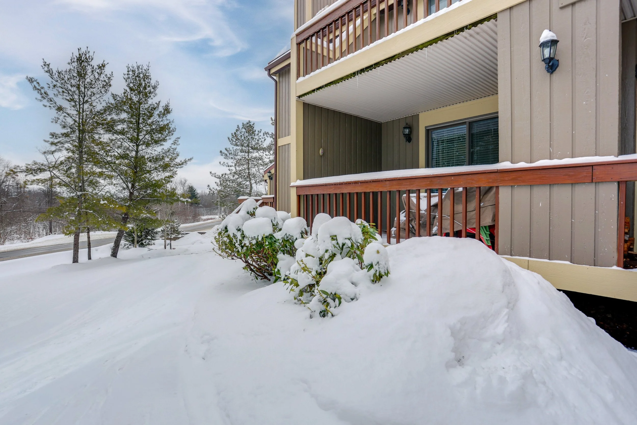 Snow-covered bushes and ground outside a multi-story residential building with brown and beige siding, wooden railings, and outdoor lights, during winter.