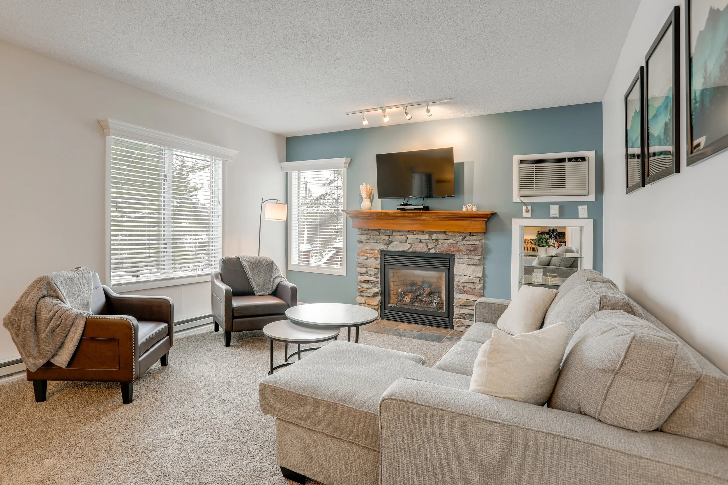 Living room with beige sofa, two black armchairs with blankets, a round coffee table, a fireplace with a wooden mantel, a mounted TV, windows with white blinds, and wall art.