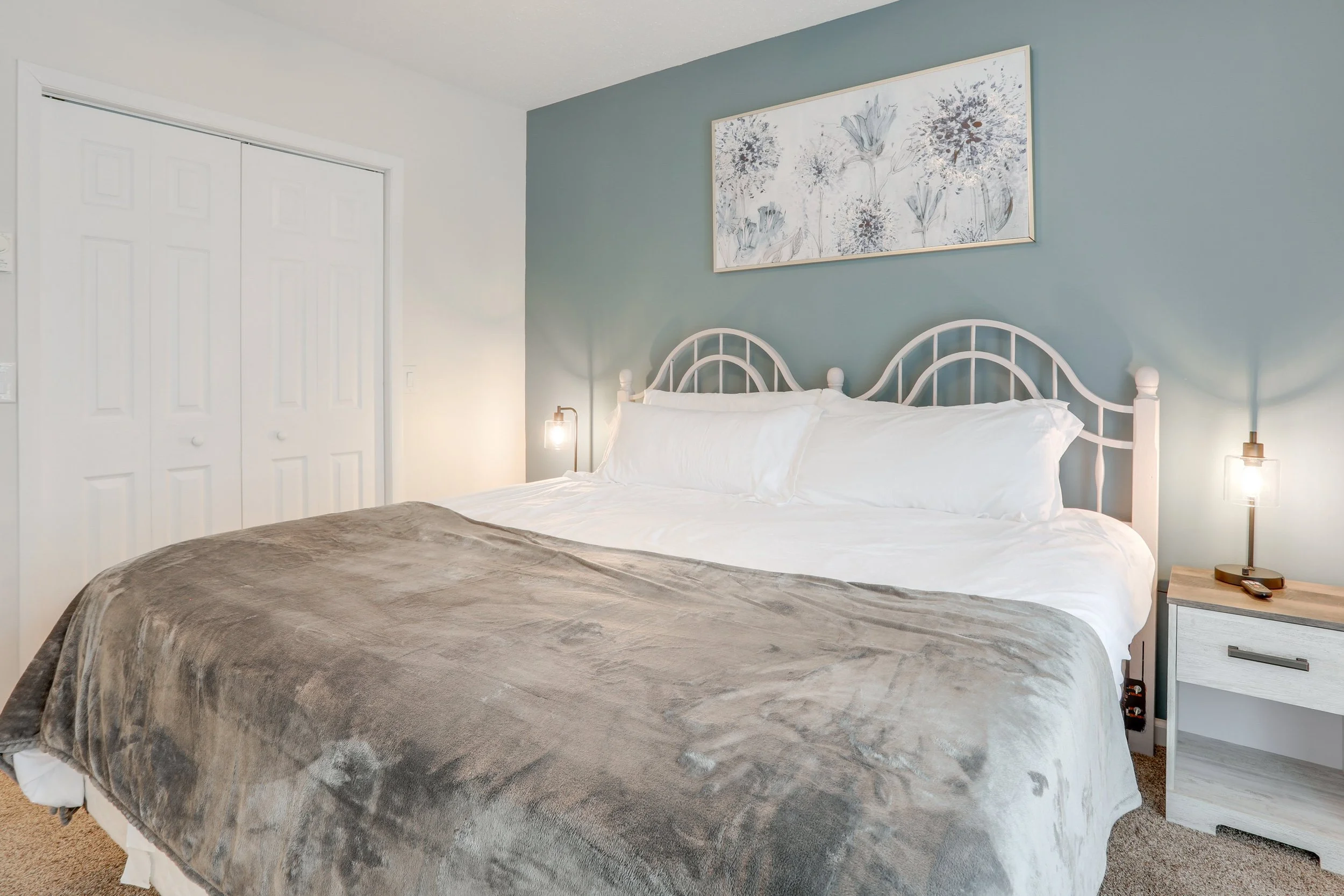 A neatly made bed with white pillows, a white headboard, and a gray blanket in a bedroom with a blue accent wall, a nightstand with a lamp and TV remote, and a framed floral artwork above the bed.