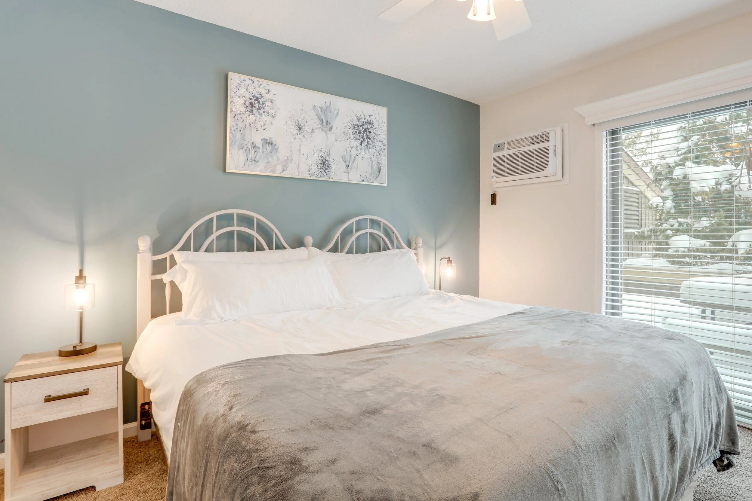 A cozy bedroom with a white metal bed frame, white bedding, and a beige blanket. There are two matching bedside lamps, a small wooden nightstand, a window with blinds, and an air conditioning unit. Artwork with floral motifs hangs on the blue accent 