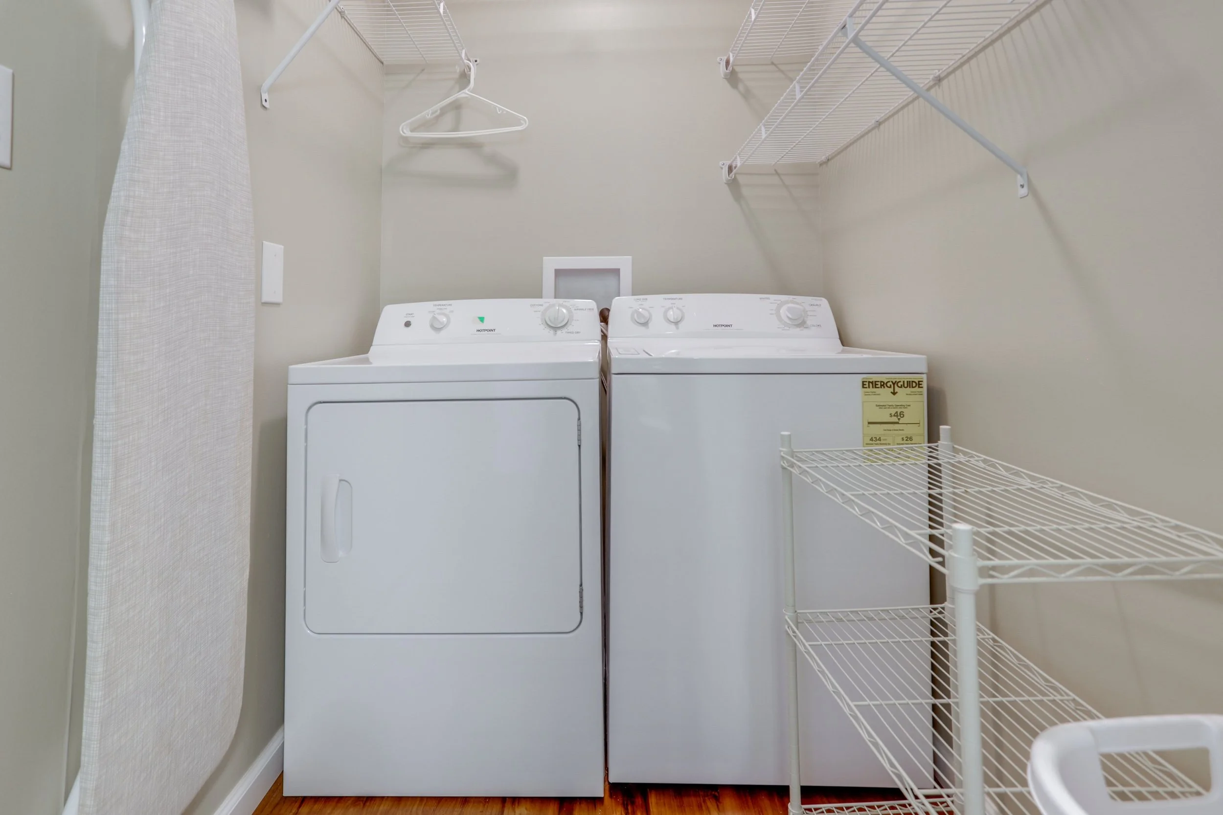 A laundry room with a white washing machine and dryer, wire shelves on the wall, and a wooden floor.