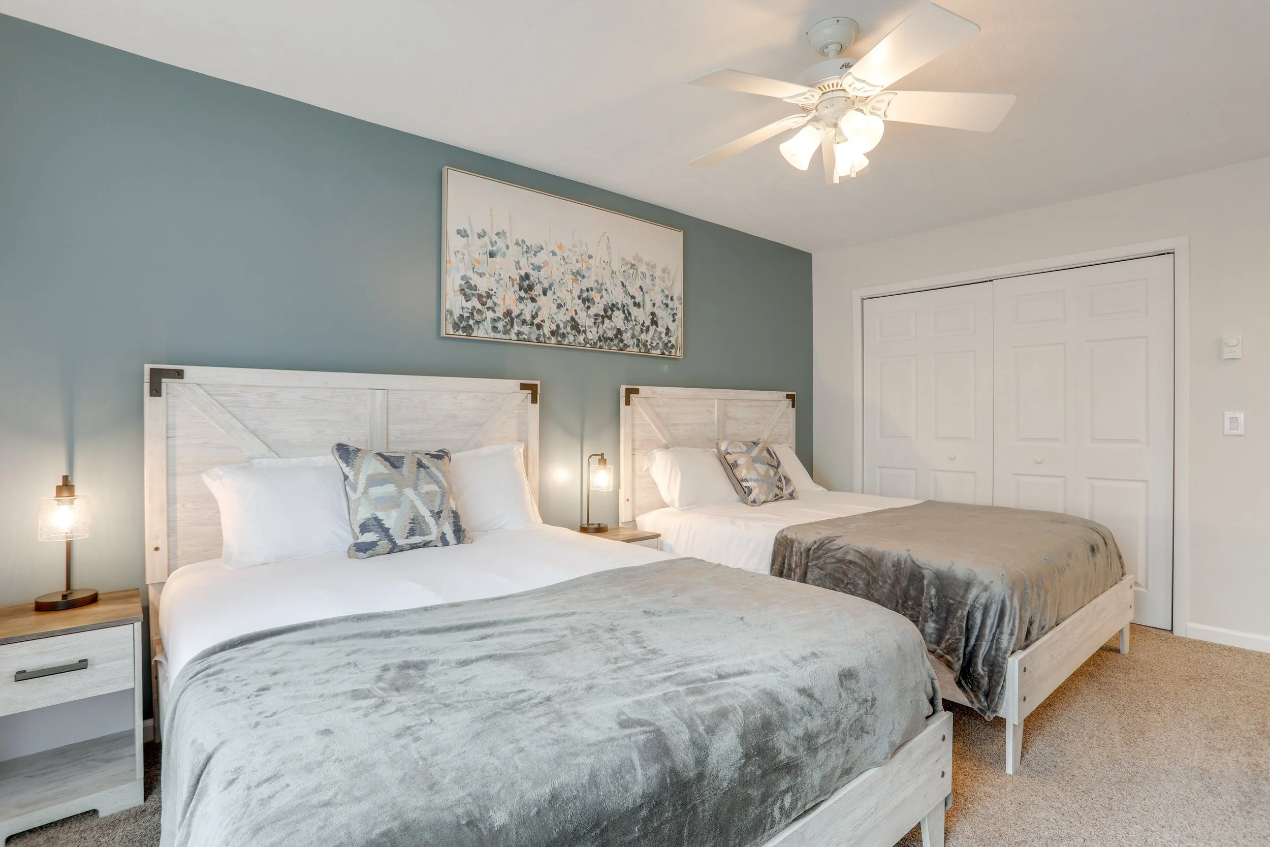 Two neatly made beds with light wood headboards and gray bedding, each with a decorative patterned pillow, in a bedroom with a blue accent wall, wall art above, and white walls and closet doors, with ceiling fan and lamps on nightstands.