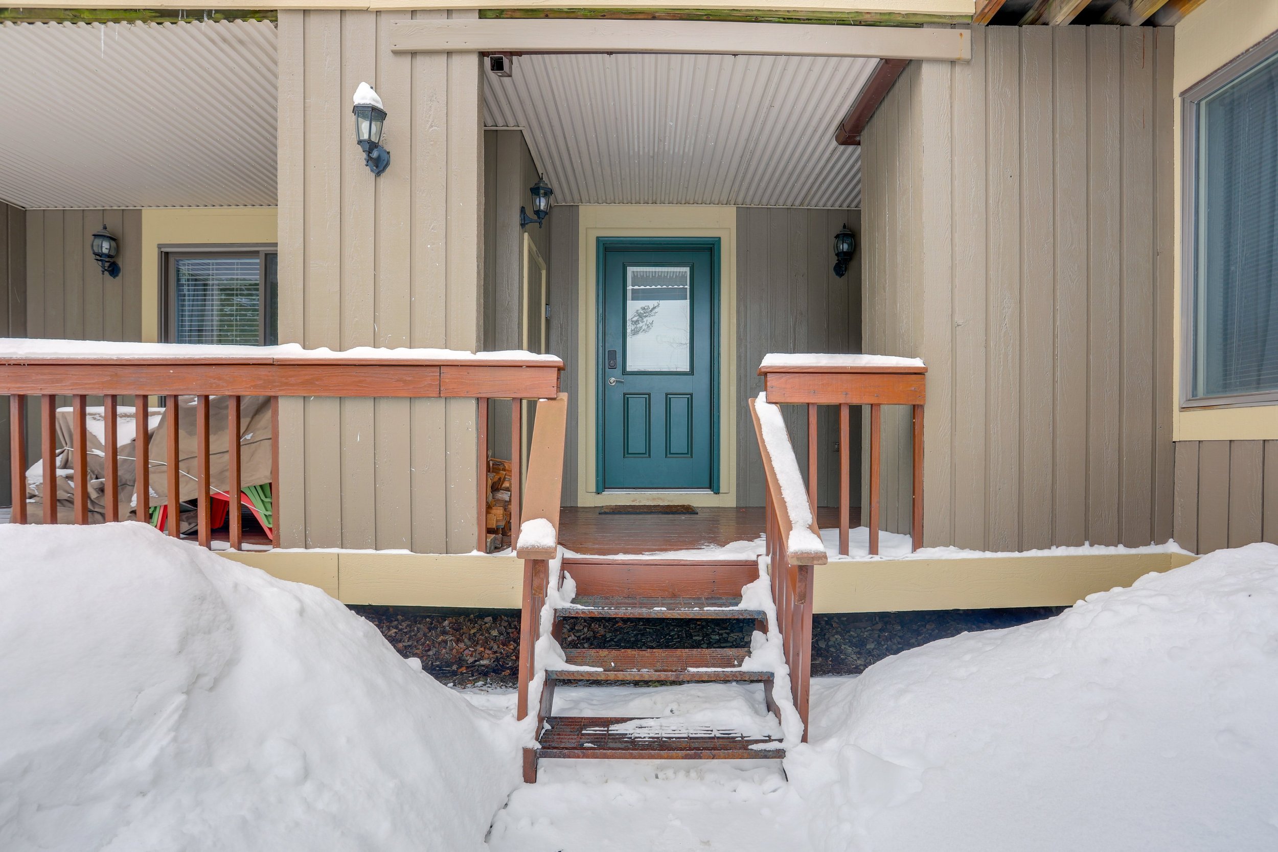 Front porch of a house with snow on the steps and railing, green door, and beige siding, with snow-covered ground in the foreground.
