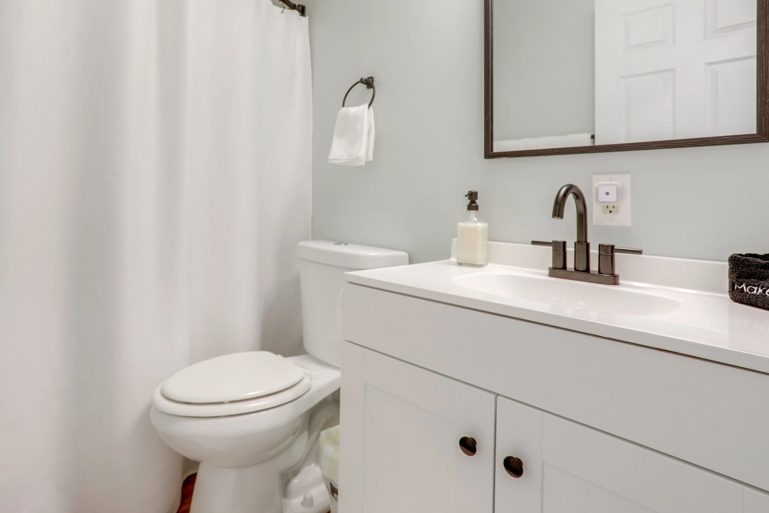 Bathroom with a toilet, white vanity with a sink, mirror, soap dispenser, towel rack with a white towel, and a black towel with embroidered text.