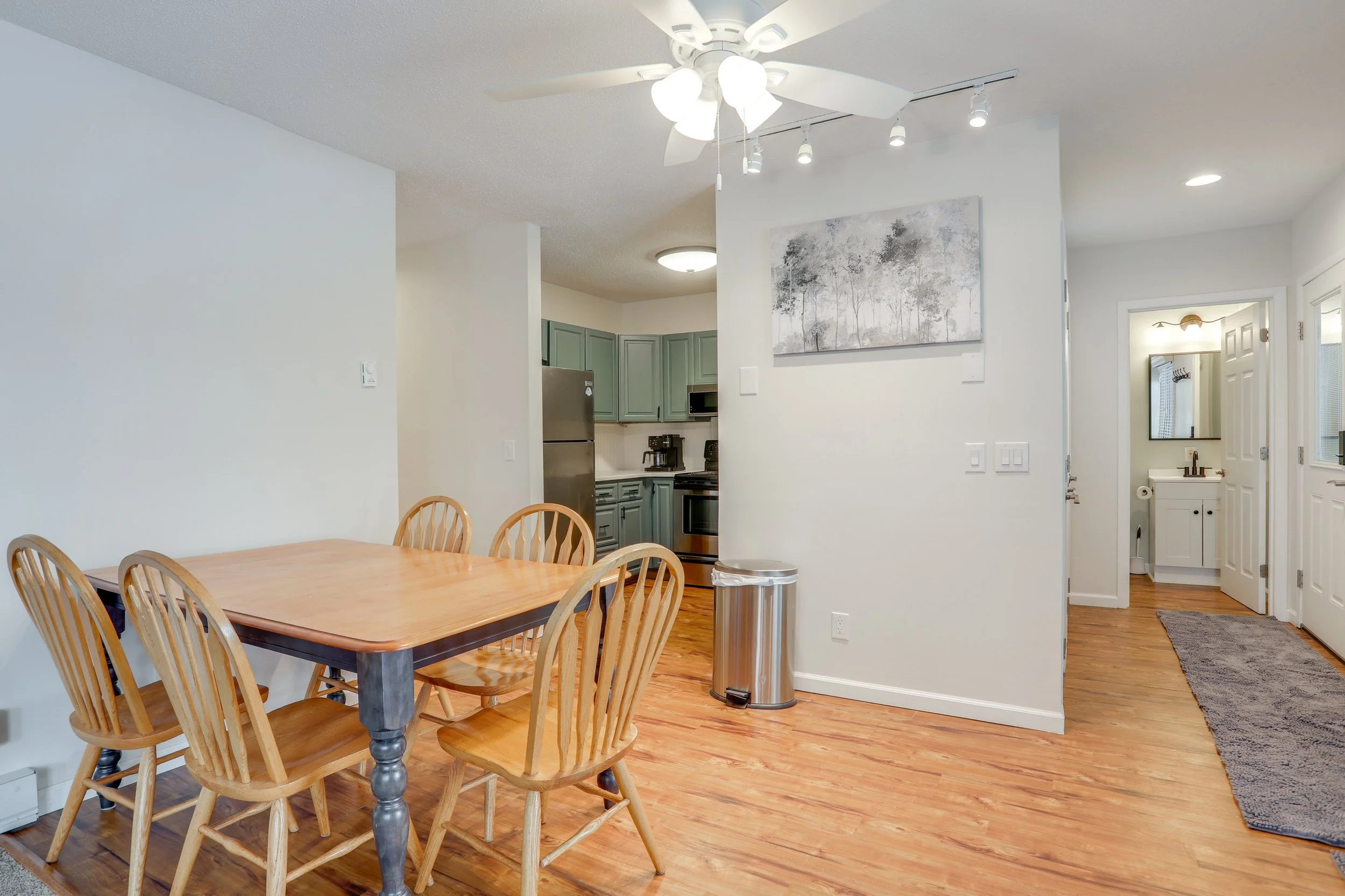 A dining area with a wooden table and six wooden chairs, hardwood flooring, a ceiling fan with lights, and a view into a kitchen with green cabinets and stainless steel appliances. There is a trash can and a hallway leading to a bathroom with a sink 