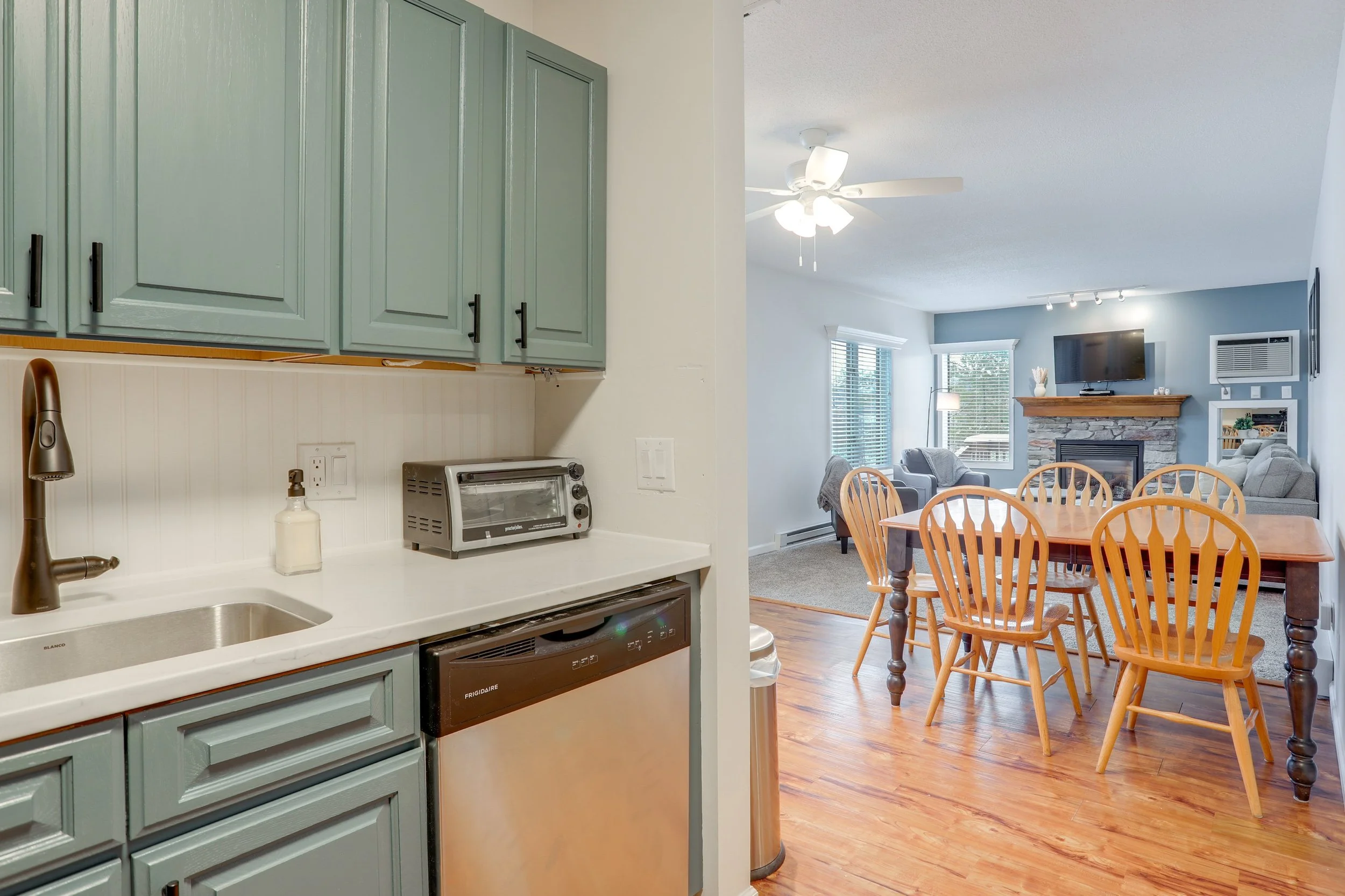 Kitchen with teal cabinets, white countertop, small toaster oven, sink, and soap dispenser. Connecting to a dining area with a wooden table and chairs, living room with gray sofa and armchairs, fireplace, TV, and windows with blinds.