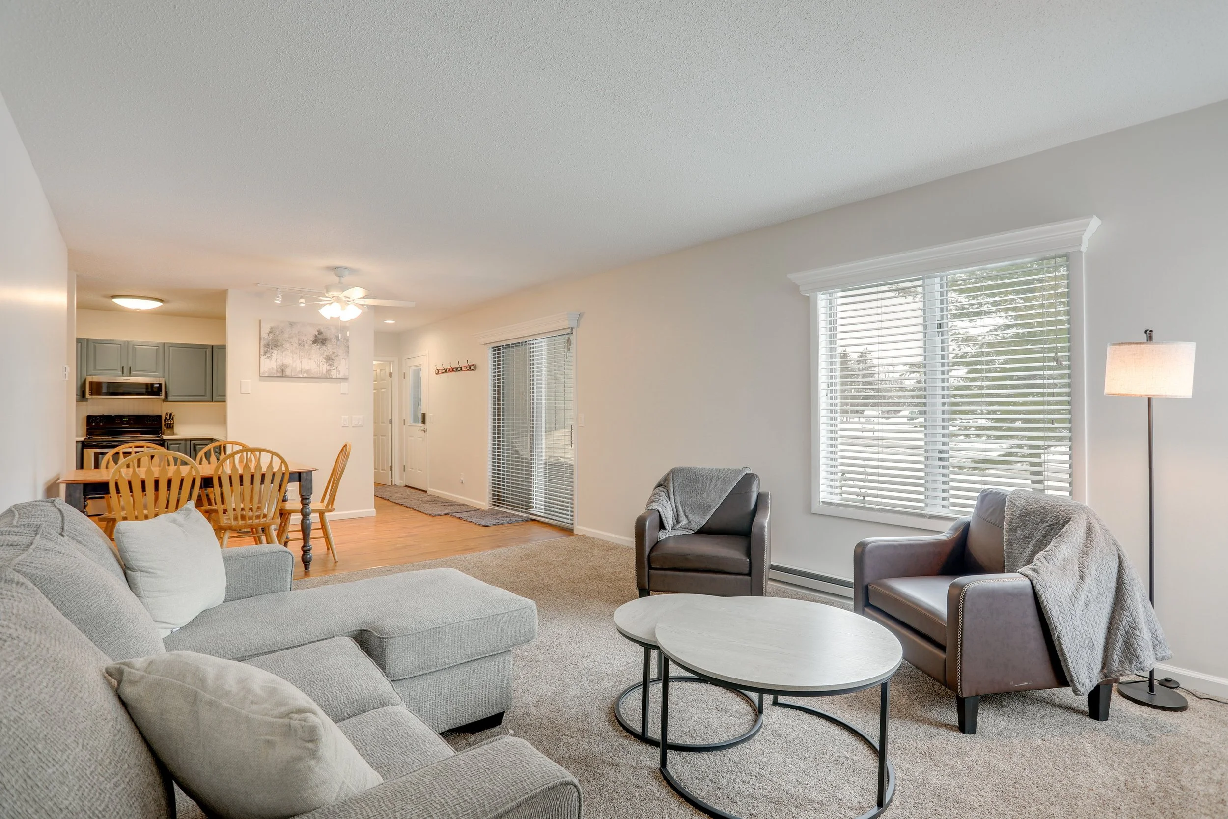 Living room with beige sofa, two armchairs, coffee table, large windows with blinds, and a floor lamp, connected to dining area with wooden table and chairs, kitchen in background with gray cabinets and stainless steel appliances.