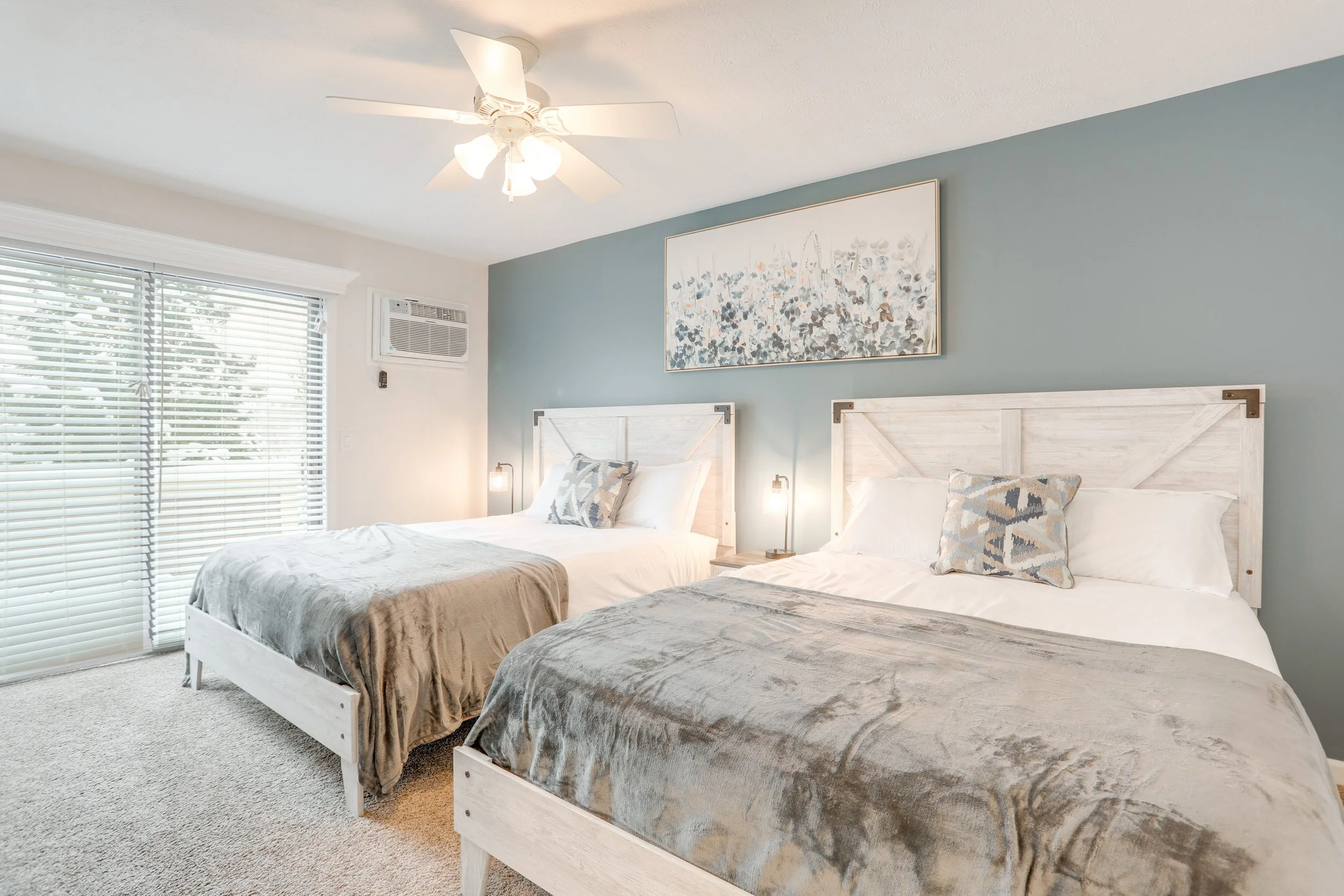 Bedroom with two beds, white wooden headboards, beige and gray bedspreads, decorative pillows, wall art, bedside lamps, window with blinds, ceiling fan, air conditioning unit.