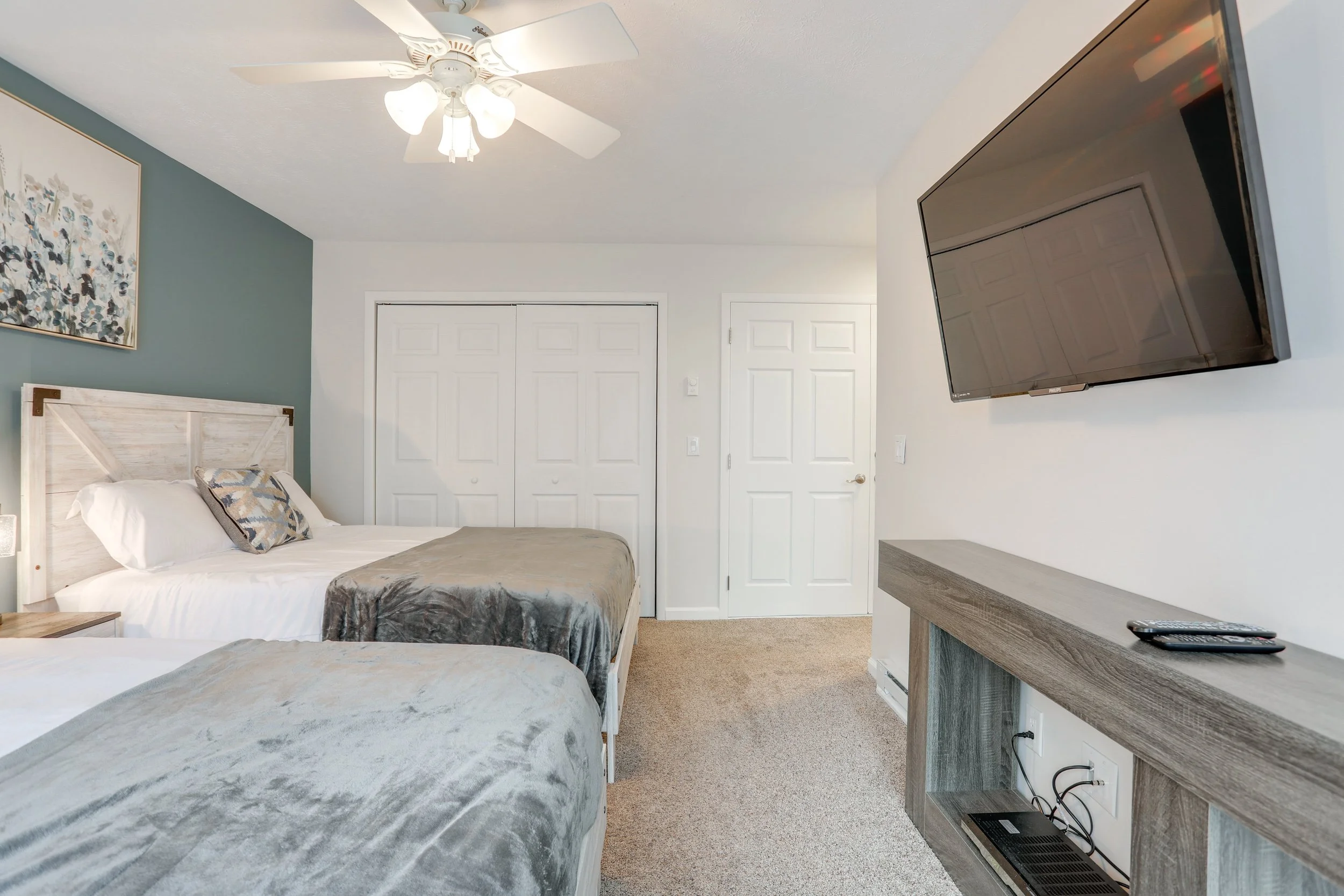A bedroom with two beds, one with a white wooden headboard, both with grey bedding and decorative pillows. A white ceiling fan with lights, a wall-mounted flat-screen TV, and a grey wooden dresser are also visible. There are closed white closet doors