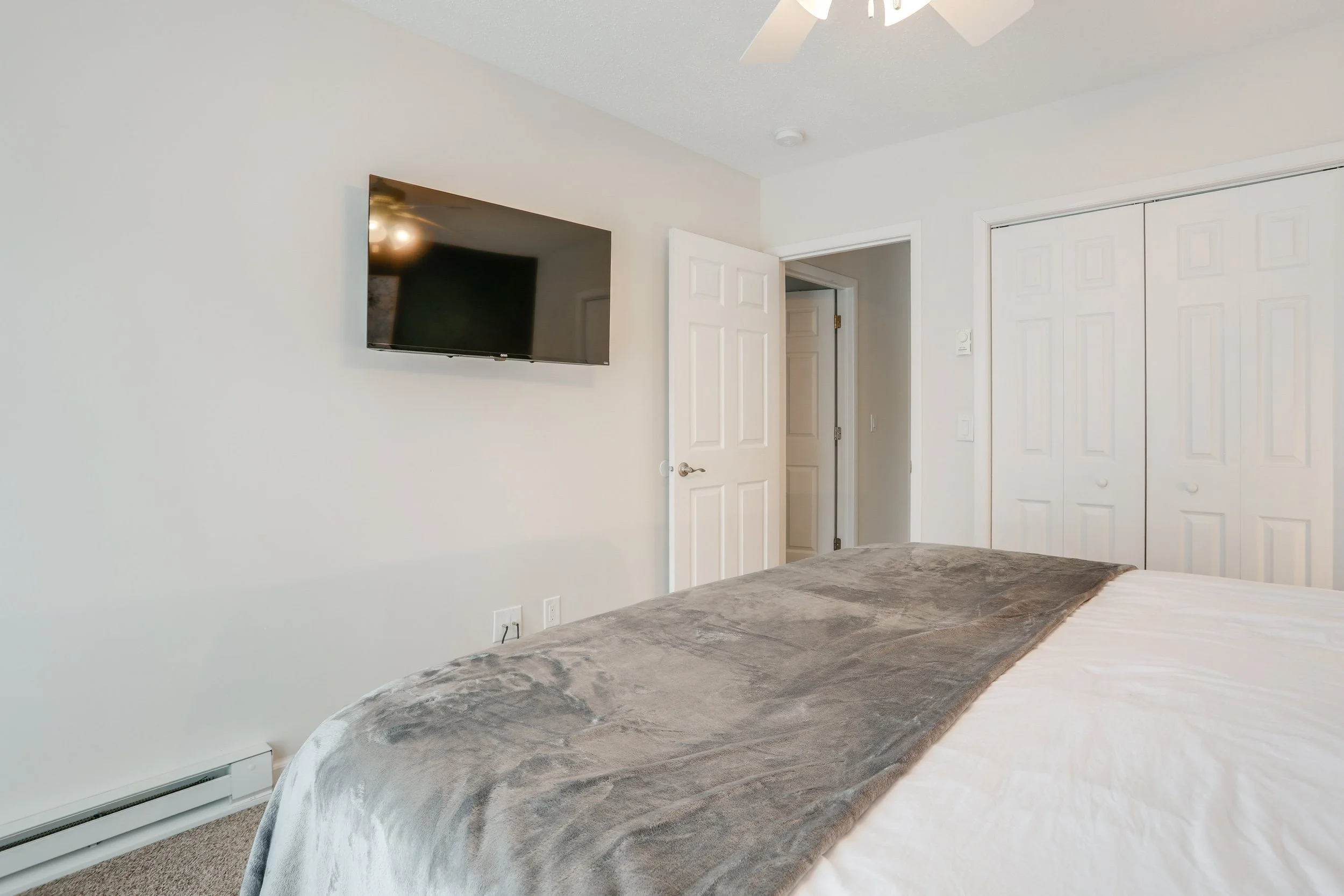 A bedroom with a flat-screen TV mounted on the wall, a bed with a gray blanket, a white door, and a closet with white bifold doors.