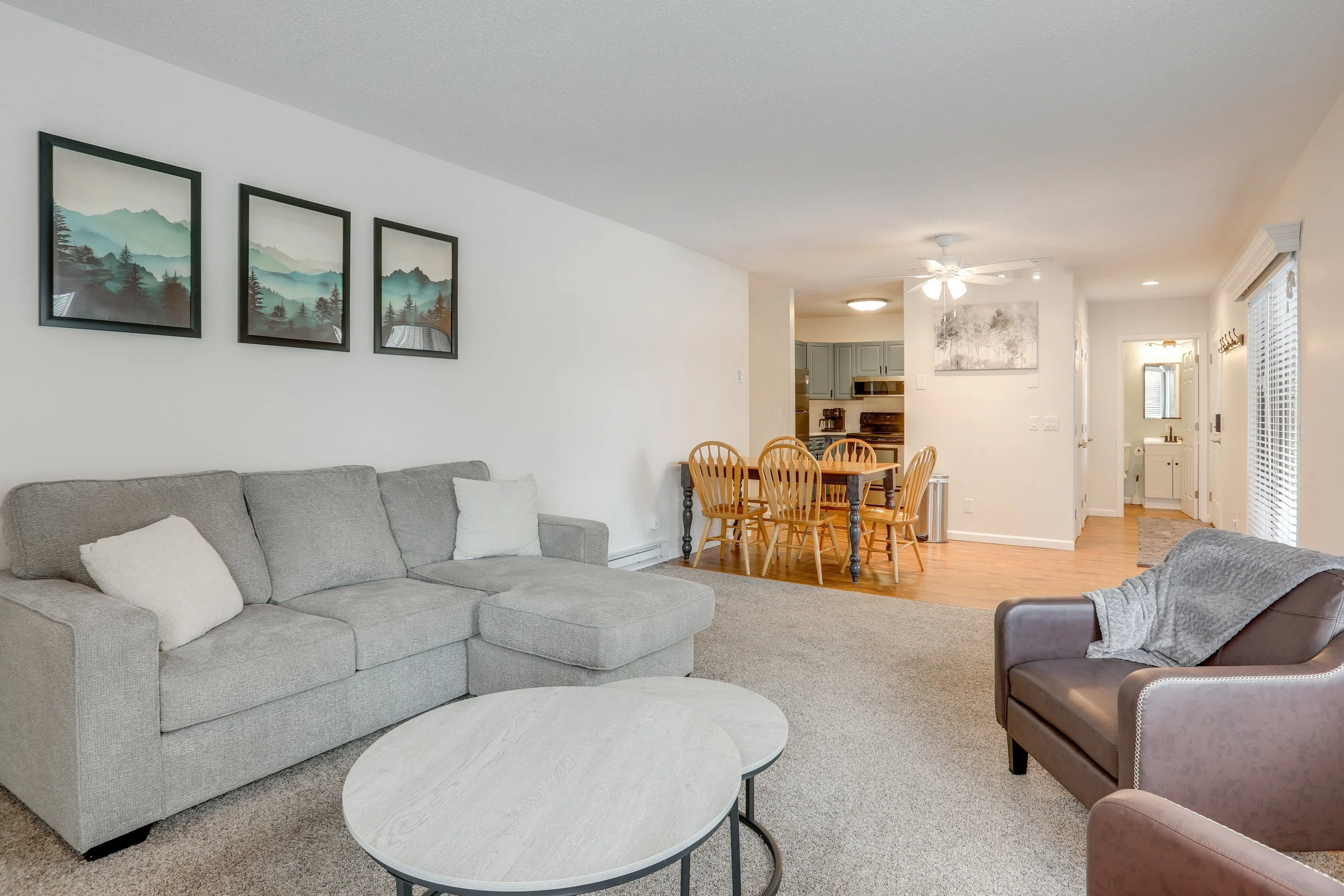 Living room with a gray sofa, a leather armchair, a wooden coffee table, and artwork of mountain landscapes on the wall. The room opens to a dining area with a wooden table and chairs and a kitchen in the background.