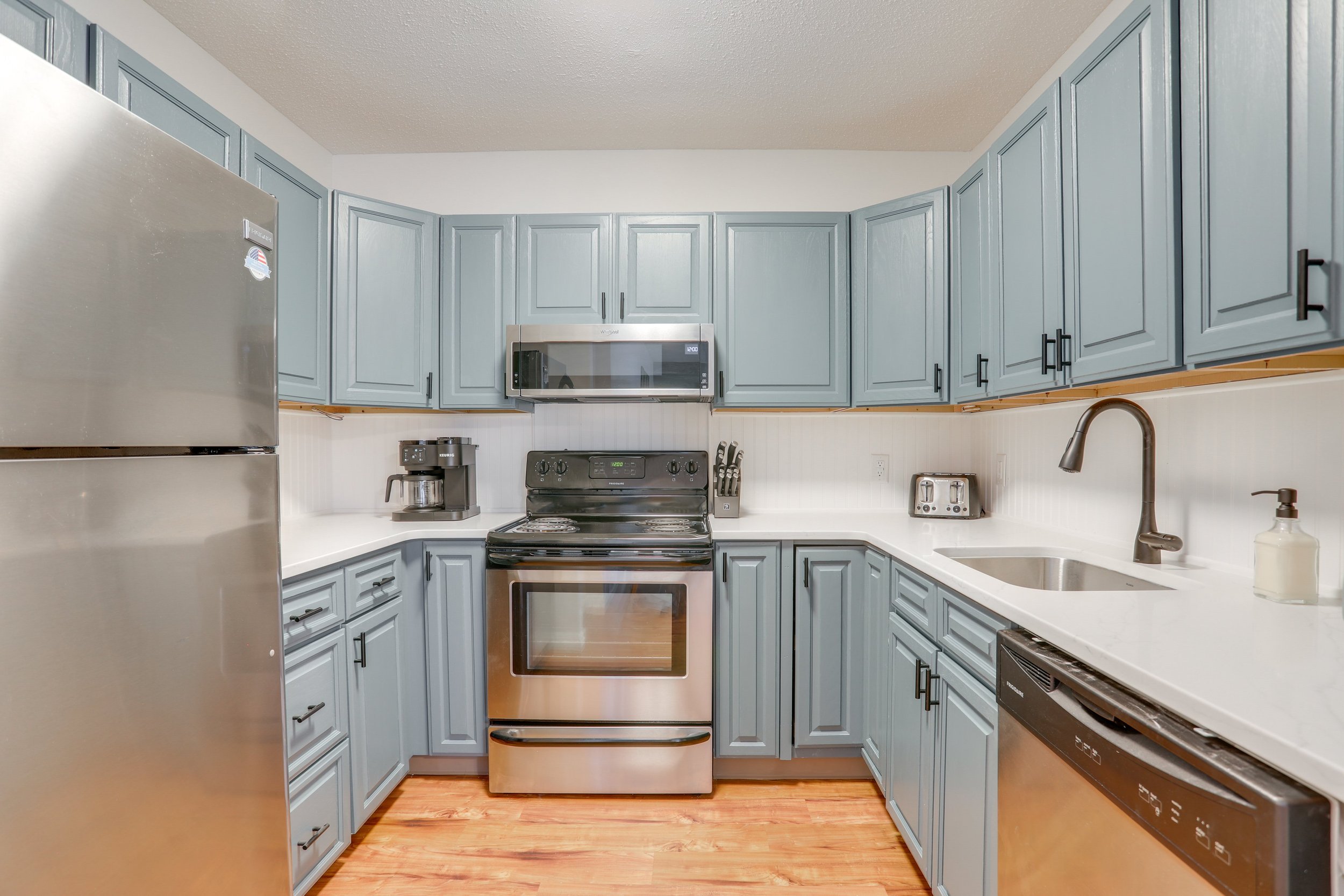 Kitchen with blue cabinets, stainless steel refrigerator, oven, microwave, coffee maker, toaster, sink, and dish soap, hardwood flooring.