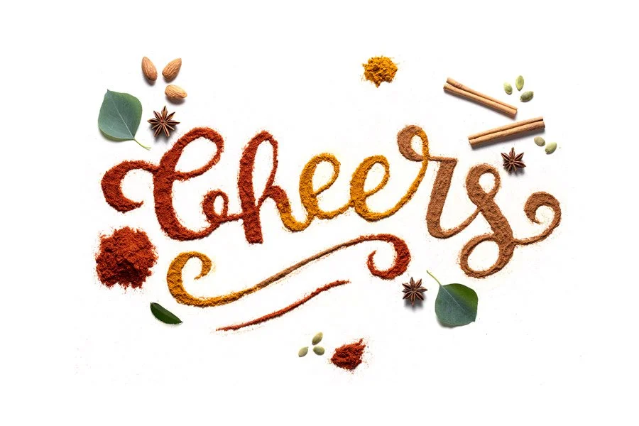 The word 'Cheers' written with various spices and herbs, including cinnamon sticks, star anise, green cardamom pods, bay leaves, and turmeric powder, arranged on a white background.
