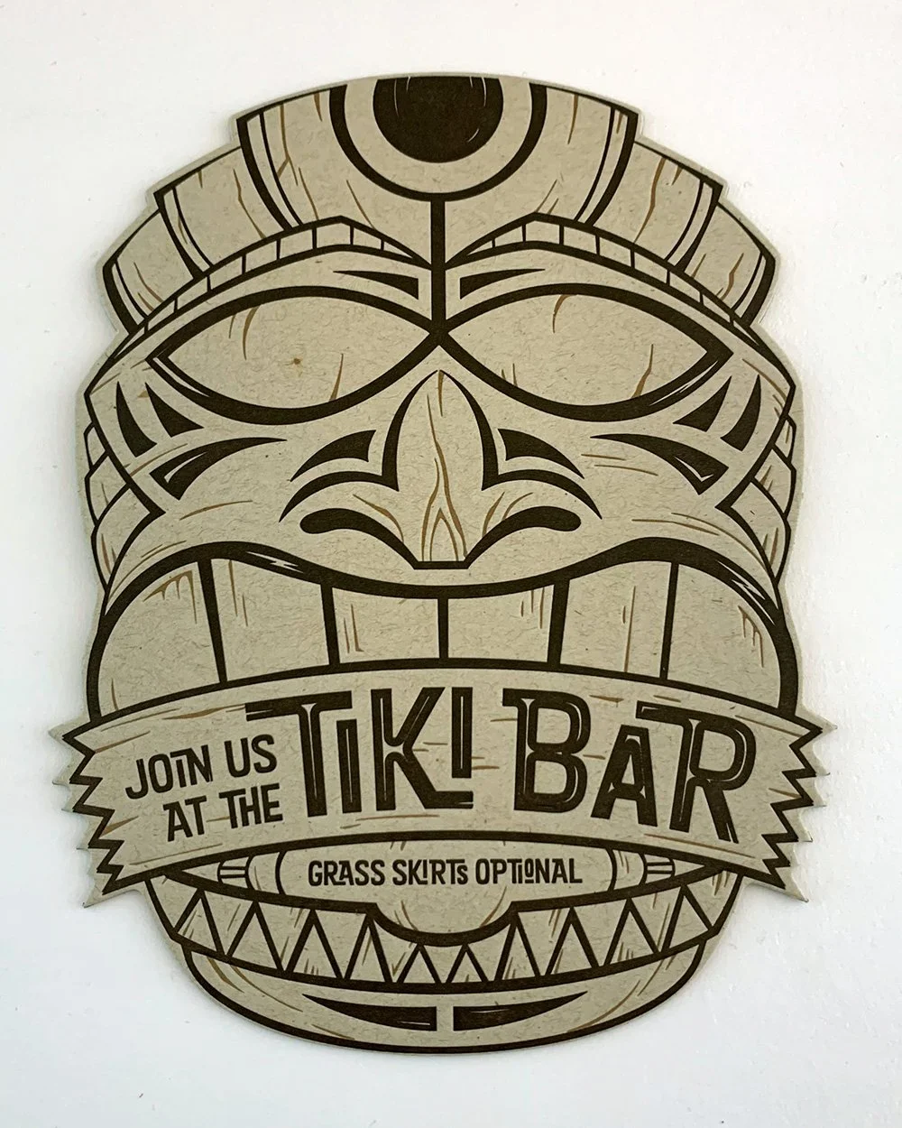 A decorative sign featuring a stylized tiki mask with bold black lines and intricate patterns. The sign promotes a tiki bar with the text "Join us at the Tiki Bar" and "Grass skirts optional."