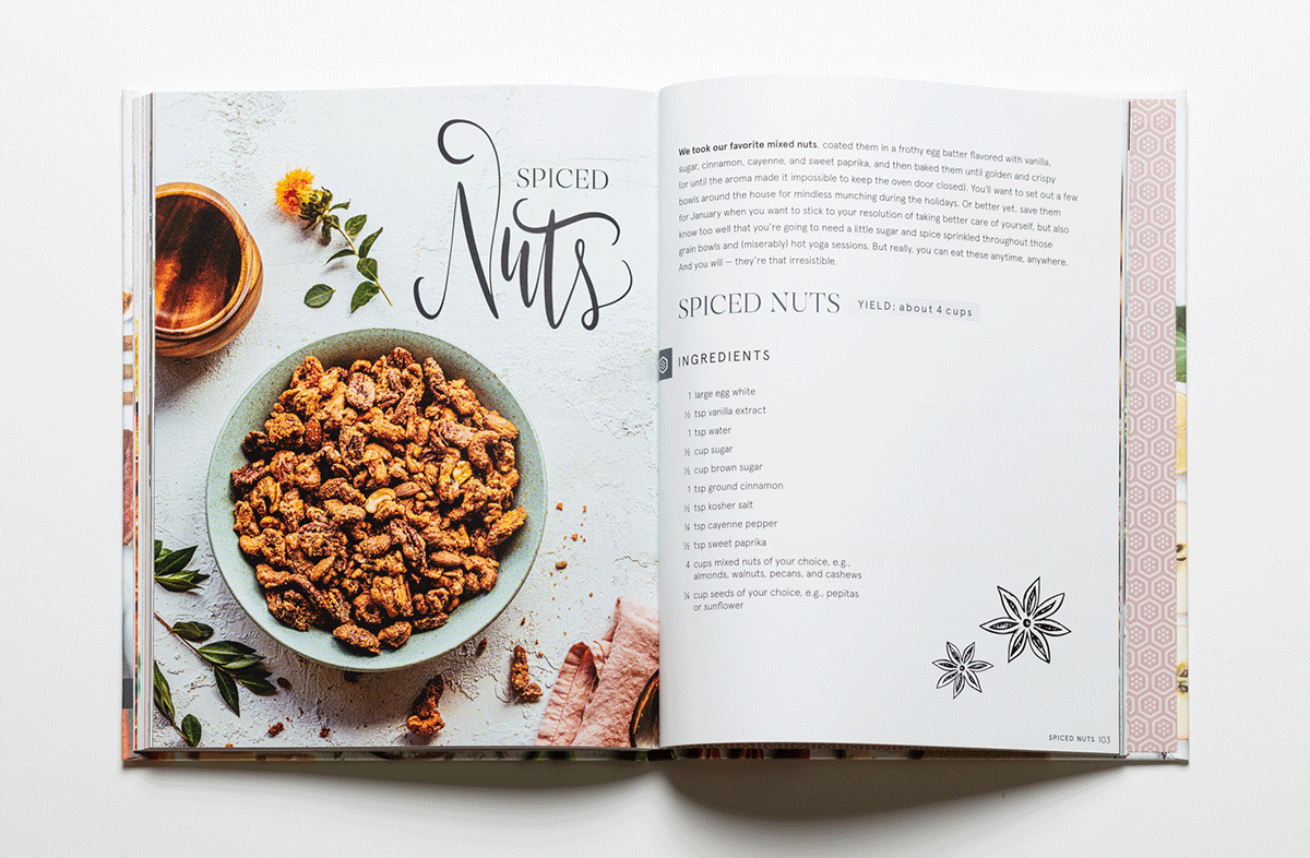 Open cookbook with a recipe for spiced nuts on the right page and a photo of spiced nuts in a bowl on the left page. The bowl contains roasted mixed nuts and there are decorative plants around.