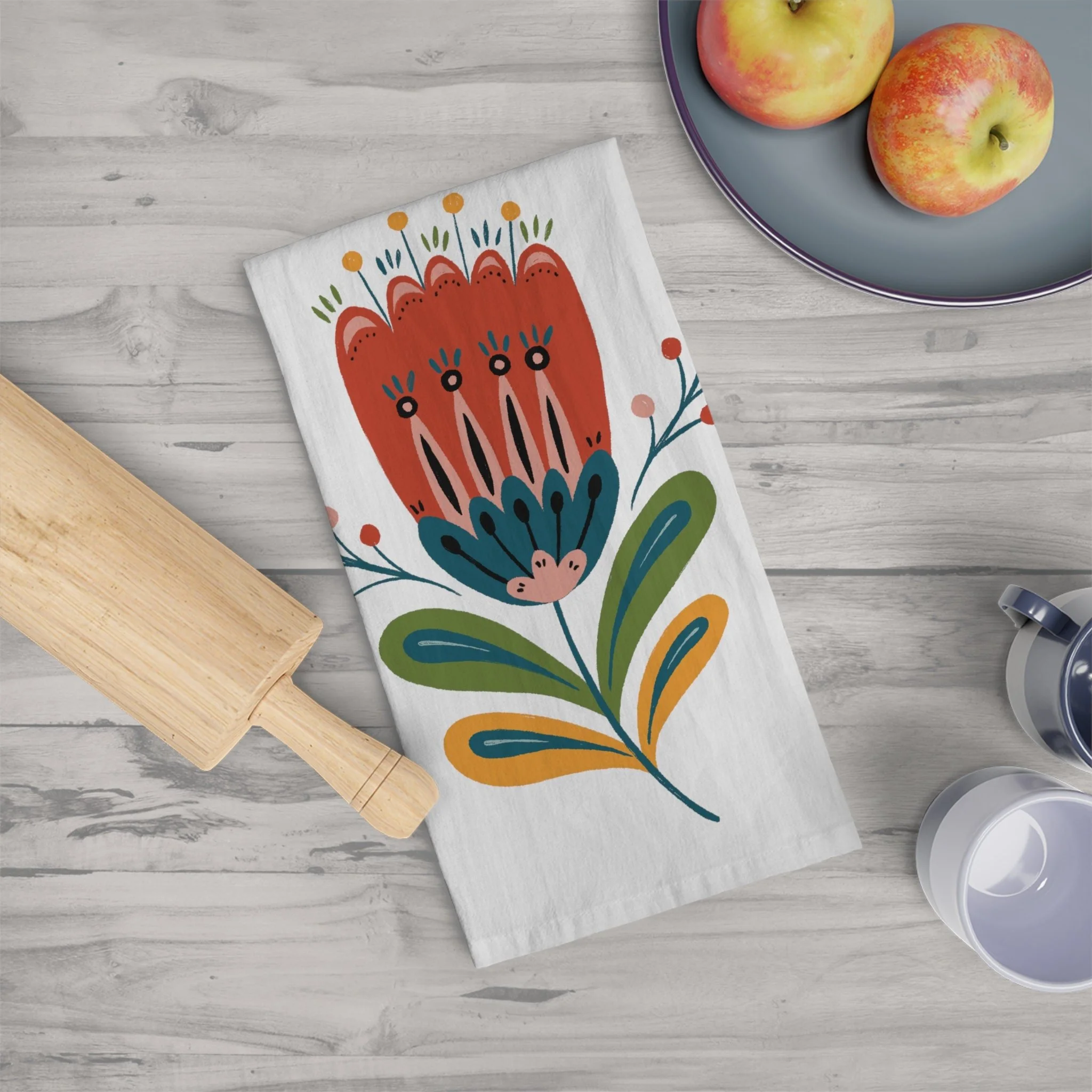 A white tea towel with a colorful, abstract folk art flower design, a gray plate with two apples, a wooden cutting board, and two cups of black coffee on a light gray wooden surface.