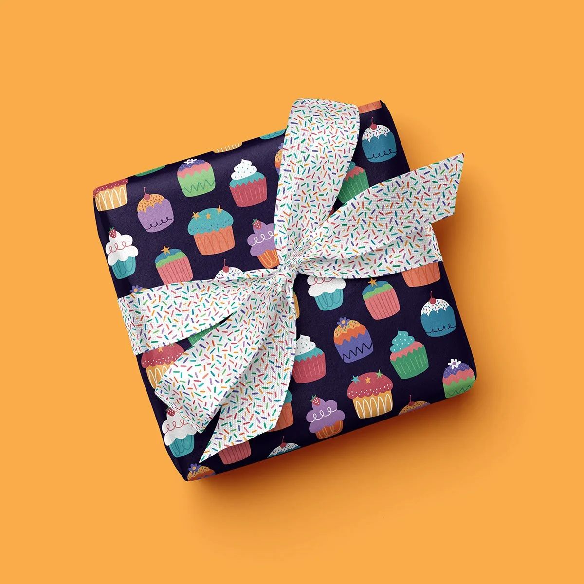 Wrapped gift box decorated with cupcake patterns and a multicolored sprinkles ribbon against an orange background.