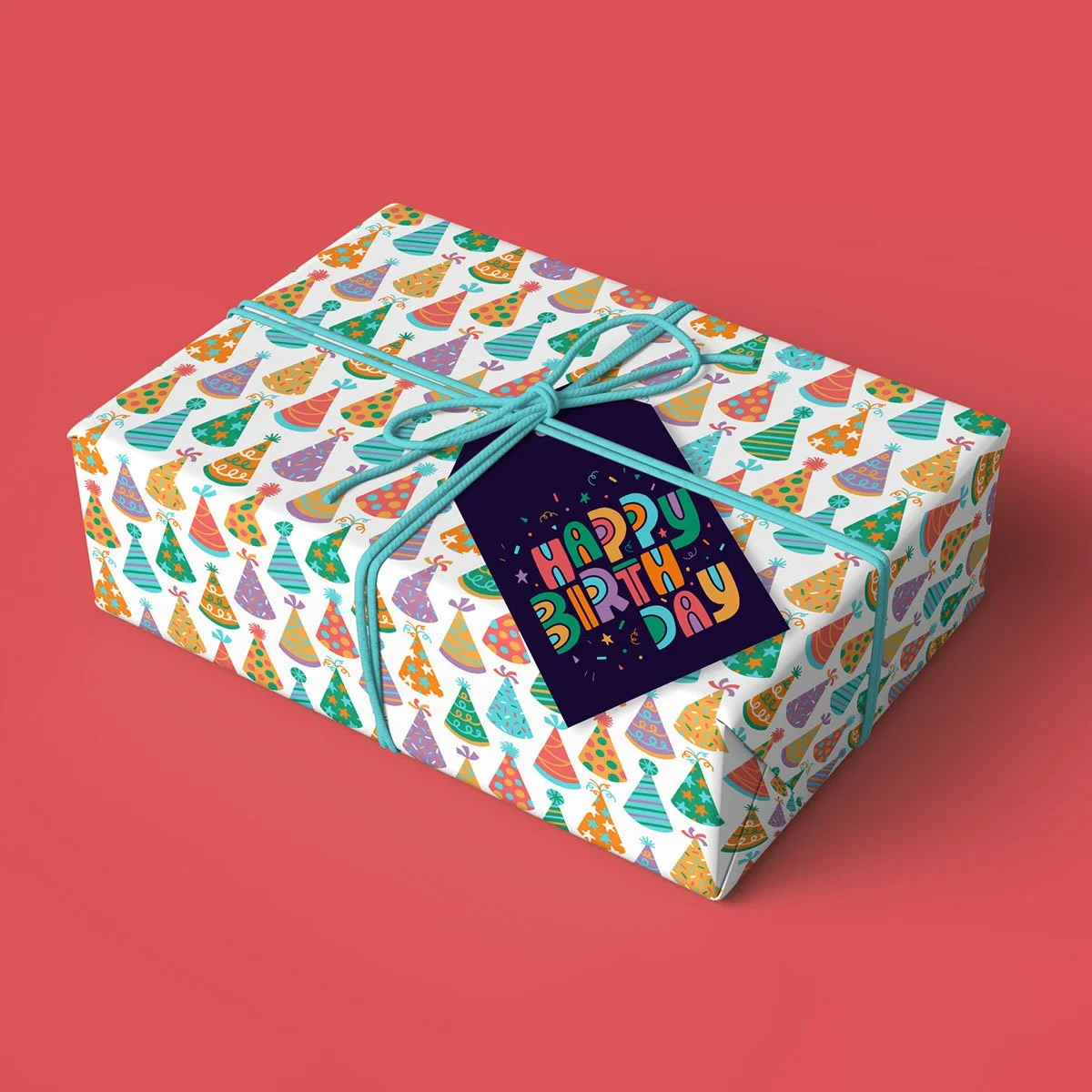 A birthday gift wrapped in party hat patterned paper, tied with blue string, with a colorful "Happy Birthday" card attached, placed on a pink surface.