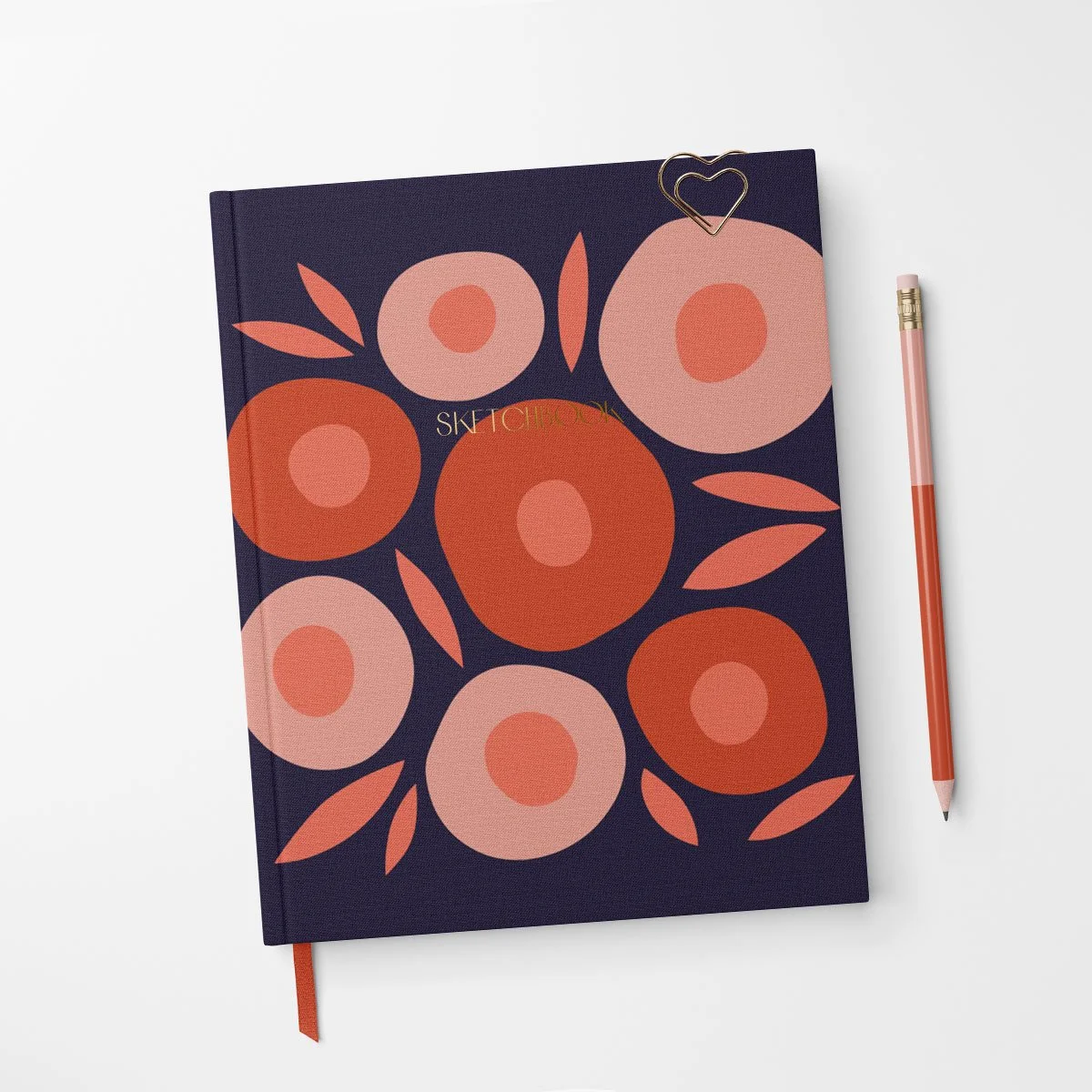 A closed notebook with a dark navy cover decorated with large pink and orange abstract flowers and leaves, with a heart-shaped paperclip attached at the top right corner, placed on a white surface alongside a pink and orange striped pen.