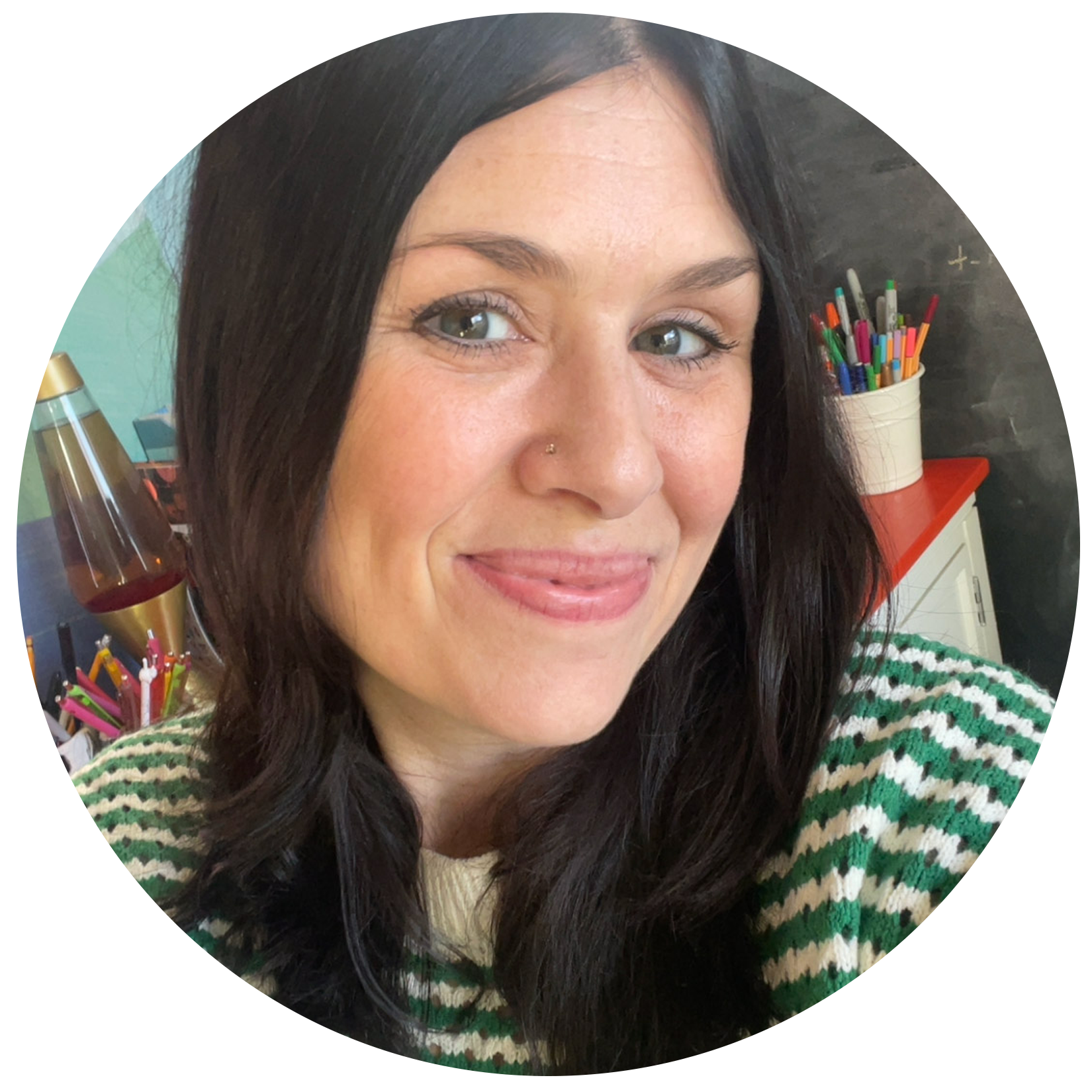 Close-up of Minneapolis artist Tina Sims with long dark hair, green eyes, and a small nose piercing, wearing a green and white striped sweater, smiling in an indoor setting with pens in a container and colorful items in the background.