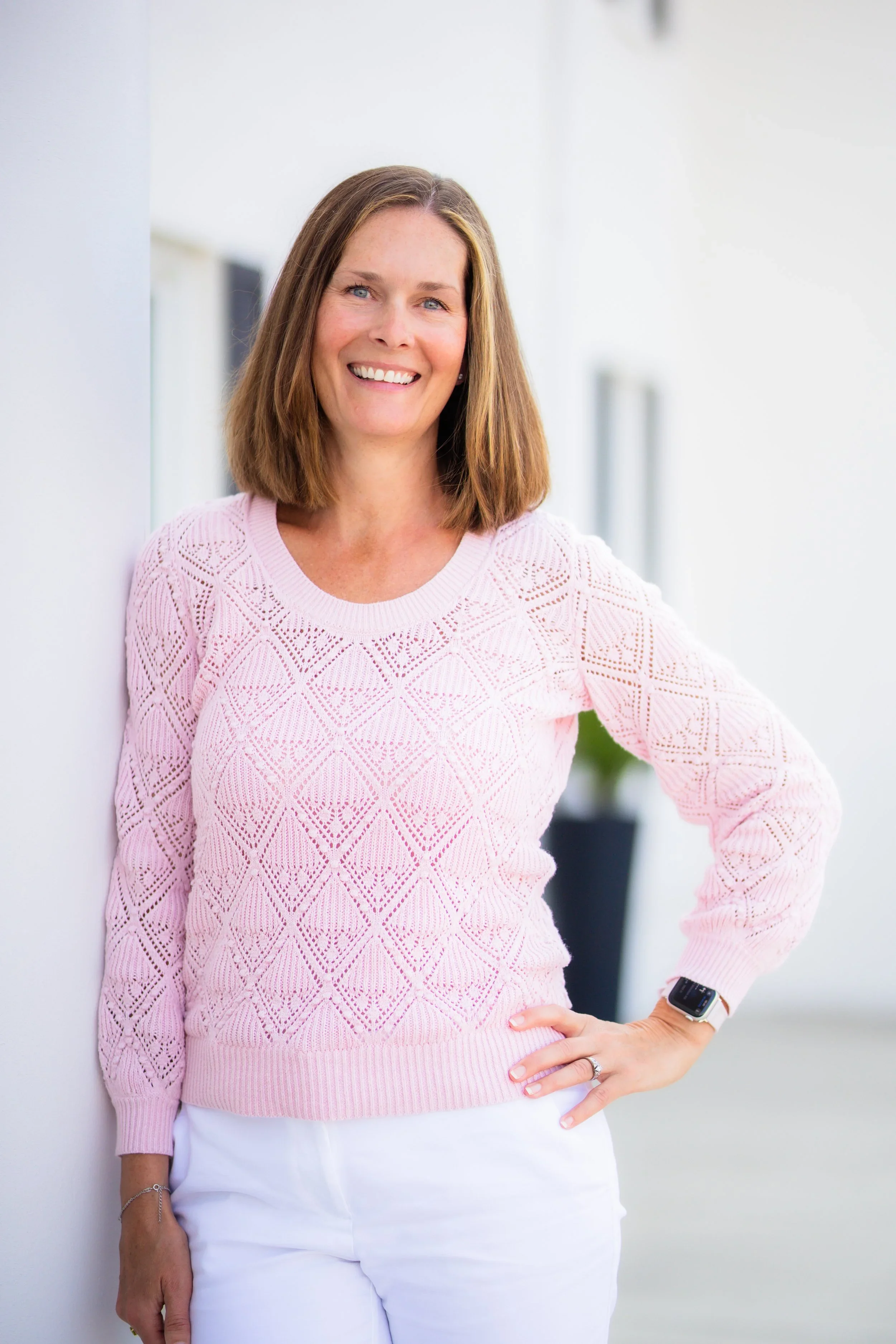 A woman with shoulder-length brown hair smiling, standing outdoors in a light pink knit sweater and white pants, with her left hand on her hip and wearing a smartwatch.