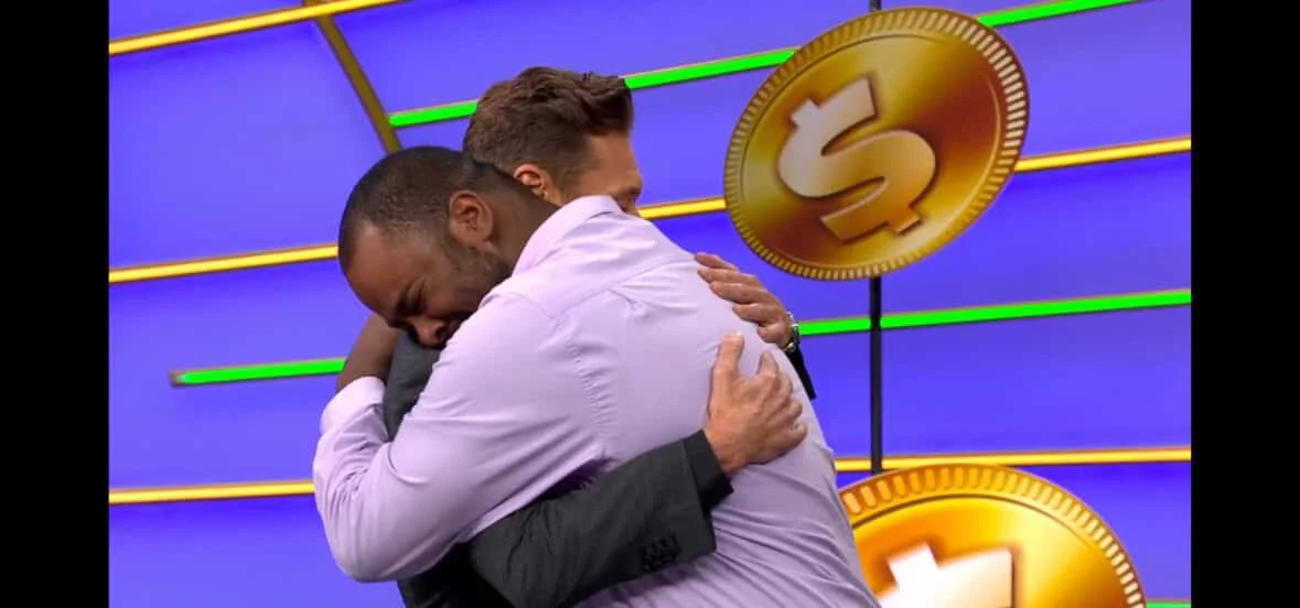 Two men hugging each other on a televised game show set, with large gold coins in the background.