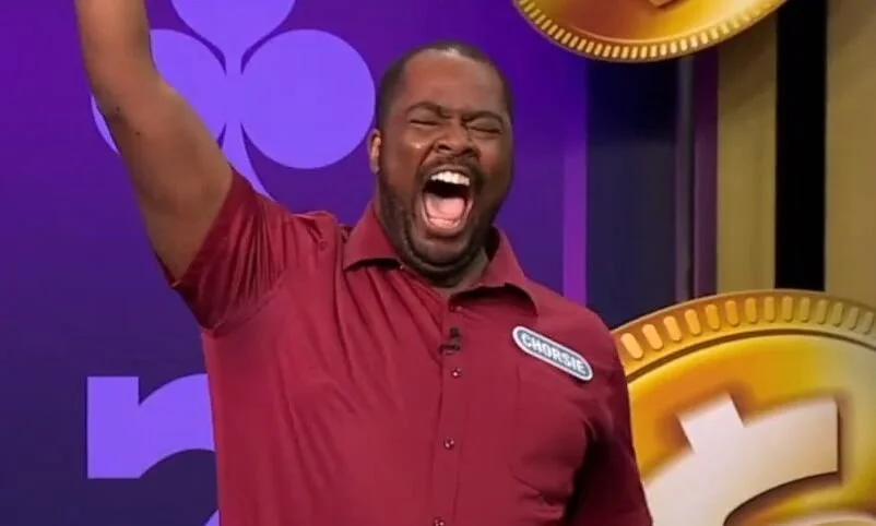 A man with a beard and mustache wearing a red shirt with a name tag, celebrating with a wide open mouth and one arm raised, on a TV game show stage with purple and gold background elements.