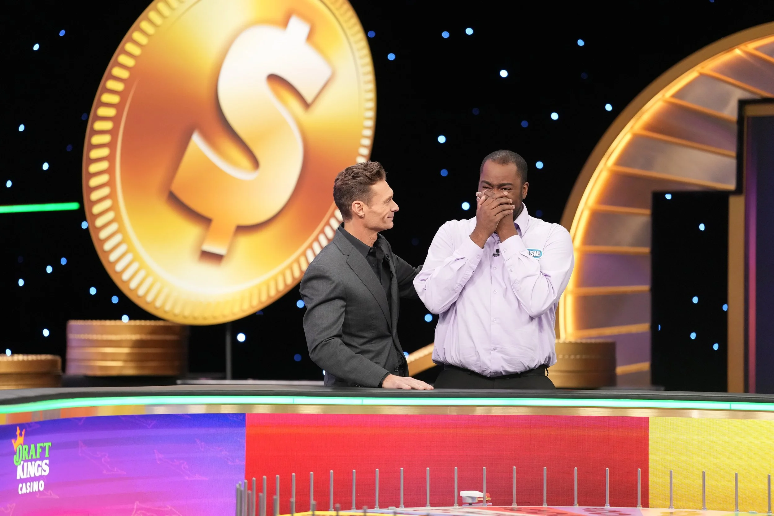 Two men standing behind a game show table, one has a hand on the other's shoulder and the other is covering his mouth and nose, with a large gold dollar sign in the background.
