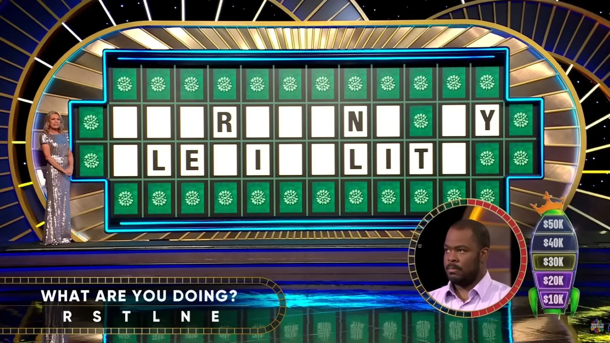 Game show screen with a puzzle featuring the phrase 'WHAT ARE YOU DOING?' with the letters 'R S T L N E' revealed. A woman stands on the left side of the stage, and a man appears on the right side in a small circular inset. The background has a gold and black circular design, and there is a wheel on the right indicating prize money up to $50K, with the $30K section highlighted.