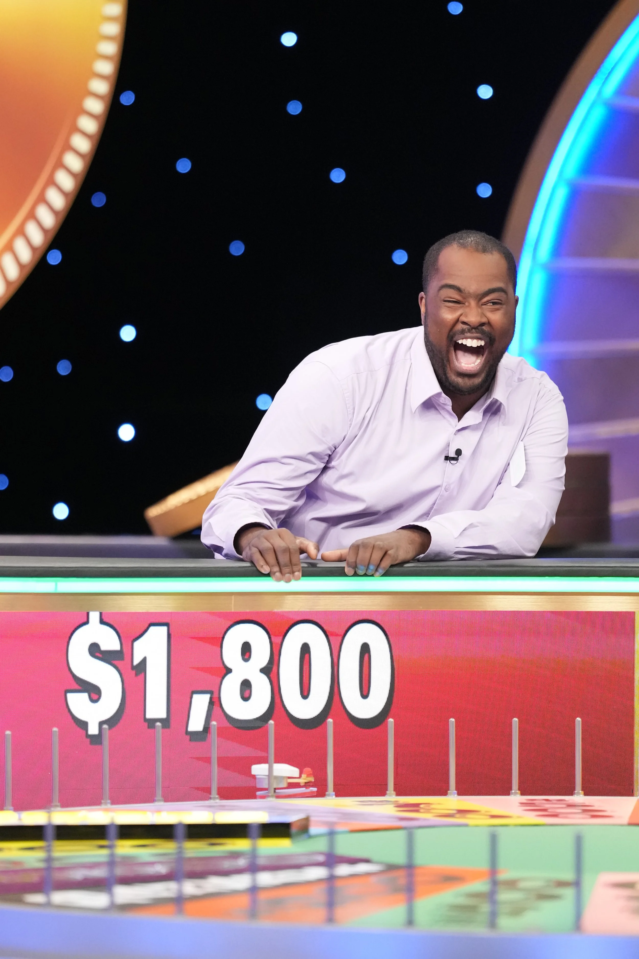 A man with a beard and mustache, dressed in a light purple shirt, laughing and leaning over a game show counter with a sign showing $1,800, on a set with a black starry background and a rainbow-colored set piece.