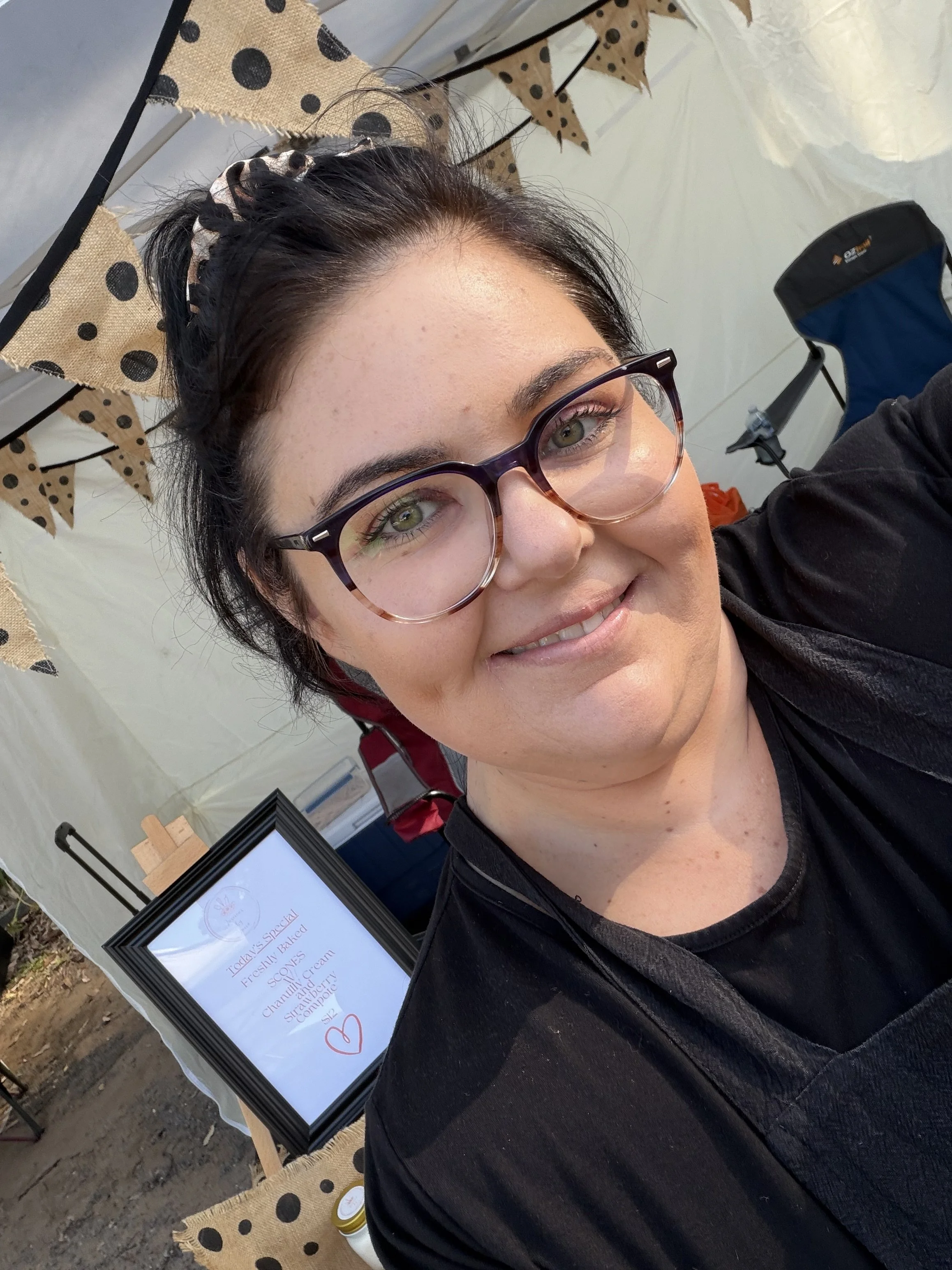 A woman with black hair, glasses, and a black shirt taking a selfie at an outdoor event. Behind her, there is a display table with a sign listing baked goods and a decorative bunting banner with polka dot flags. In the background, there is a white tent and a blue folding chair.