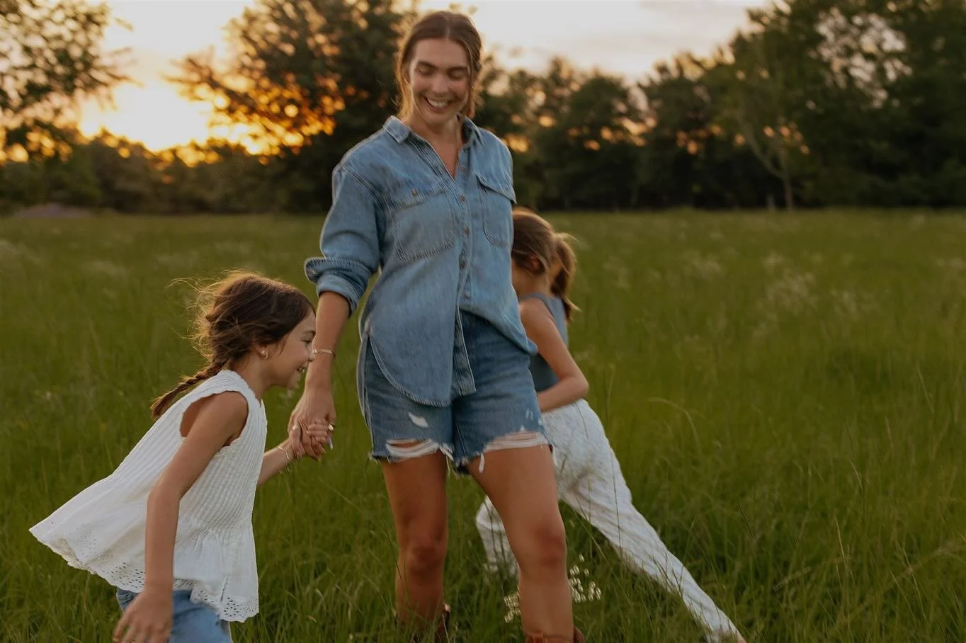 A woman holding hands with two young girls while walking through a grassy field at sunset.