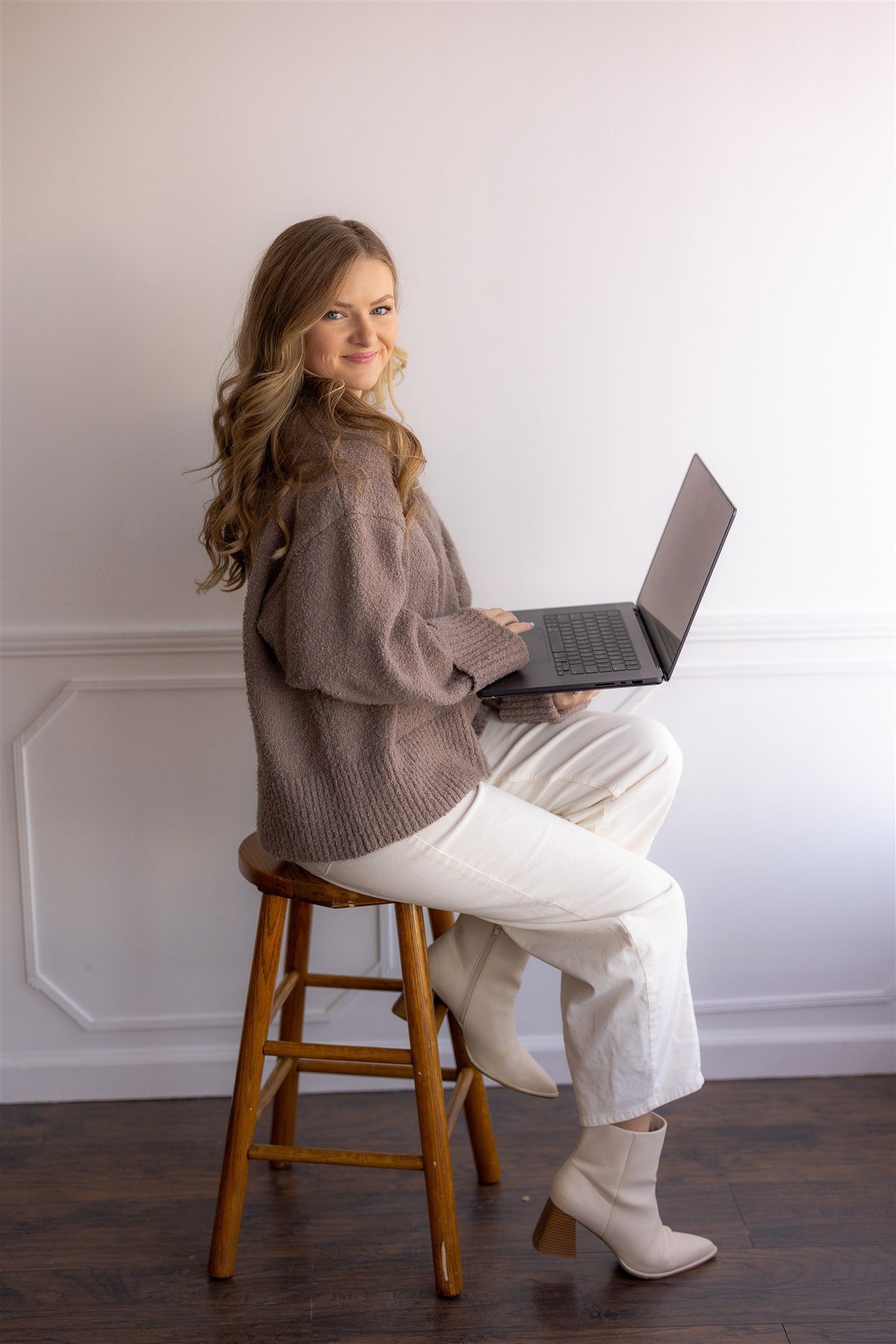Cierra Burch, Creative Brand Strategist, sitting on a wooden stool, holding a laptop on her lap, wearing a brown sweater, white pants, and beige high-heeled boots, looking at the camera with a slight smile, in front of a plain wall.