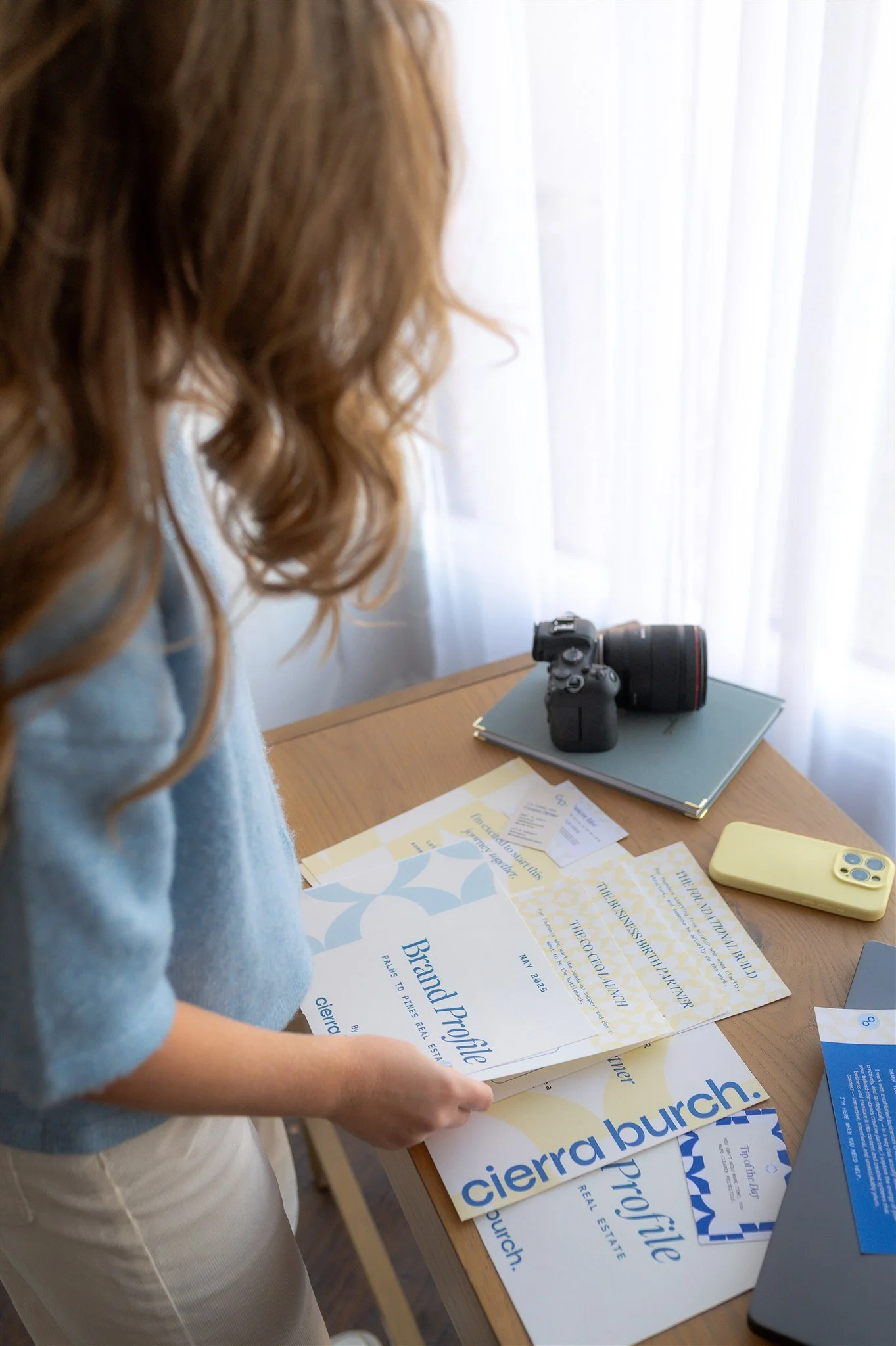 Cierra Burch, Creative Brand Strategist wearing a blue sweater and beige pants standing at a desk, looking at brochures and documents, with a camera, notebook, yellow phone, and laptop on the desk, and sheer white curtains in the background.