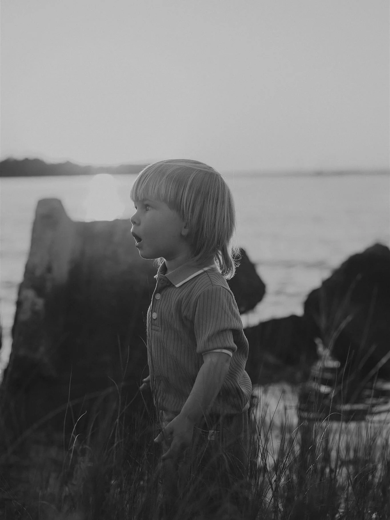 A young boy with light-colored hair standing outdoors near the water, surrounded by rocks and grass, looking to his left with an open mouth.