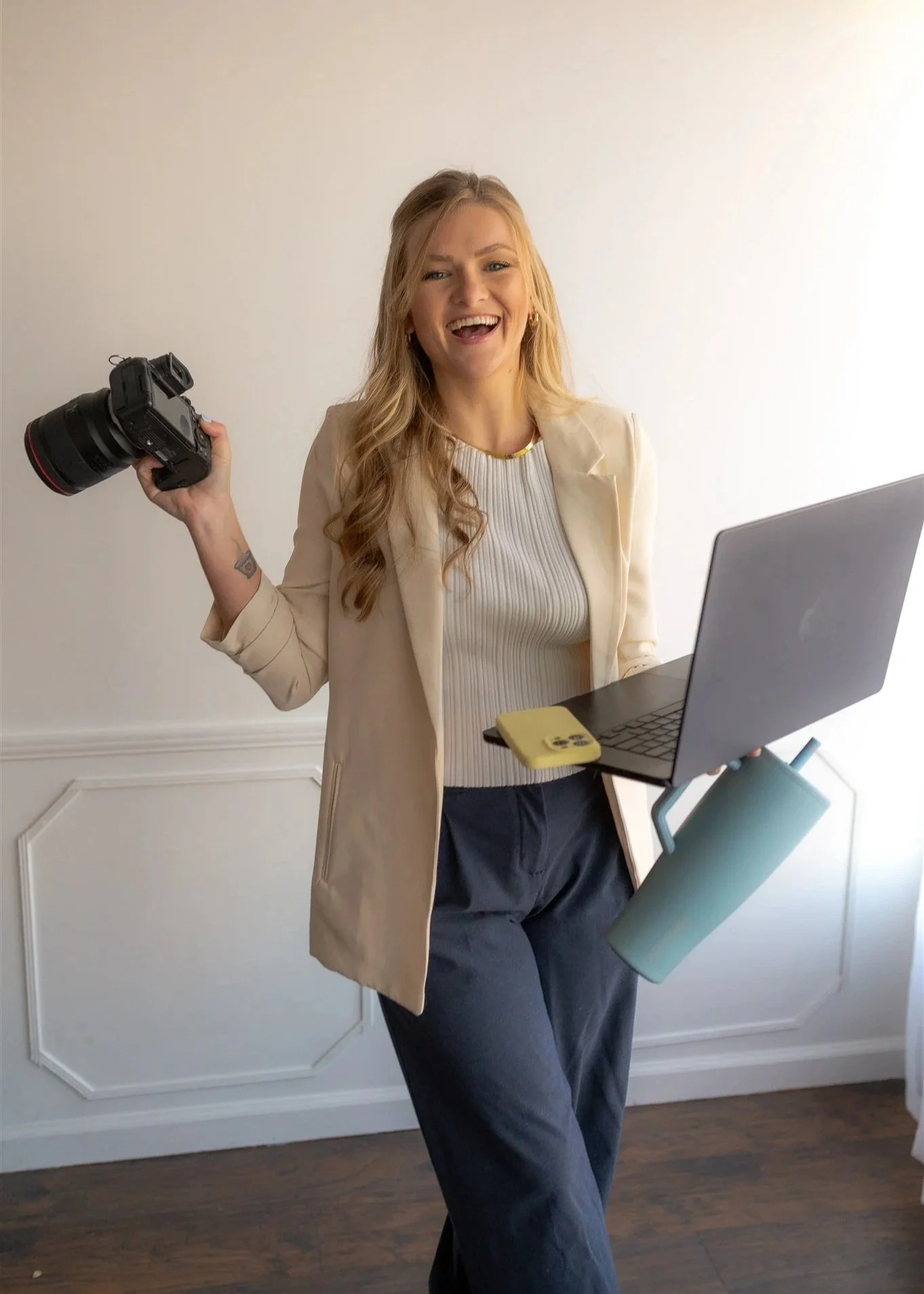 Cierra Burch, Creative Brand Strategist, smiling and holding a camera in one hand and a laptop with a paper towel roll hanging from it in the other, standing in a room with white walls and wooden floor.