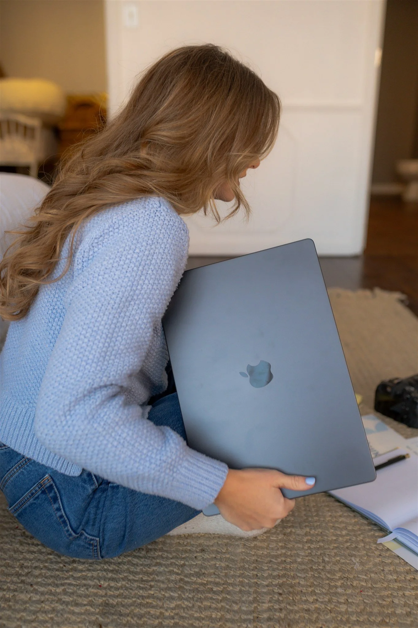 Cierra Burch, Creative Brand Strategist, holding a closed silver MacBook laptop, sitting on a carpeted floor in a room with a bed and a white dresser in the background, with papers and a camera nearby.