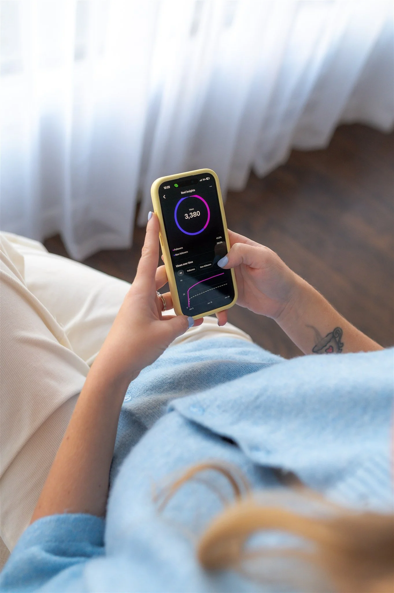 A person sitting on a couch using a smartphone to check their social media analytics, with a curtain and wooden floor in the background.