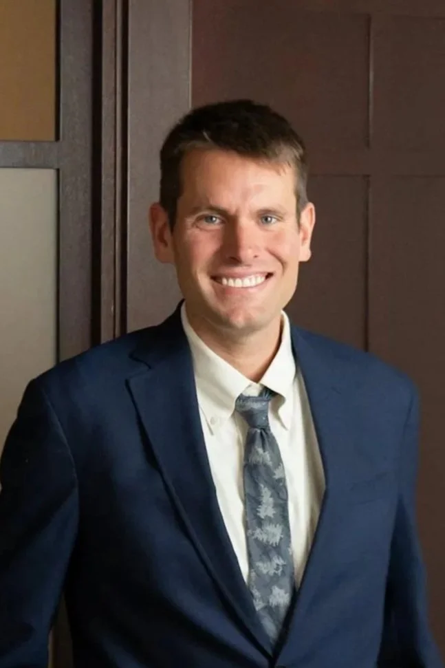 A man in a dark blue suit, white shirt, and patterned gray tie smiling and standing indoors against a wooden wall.