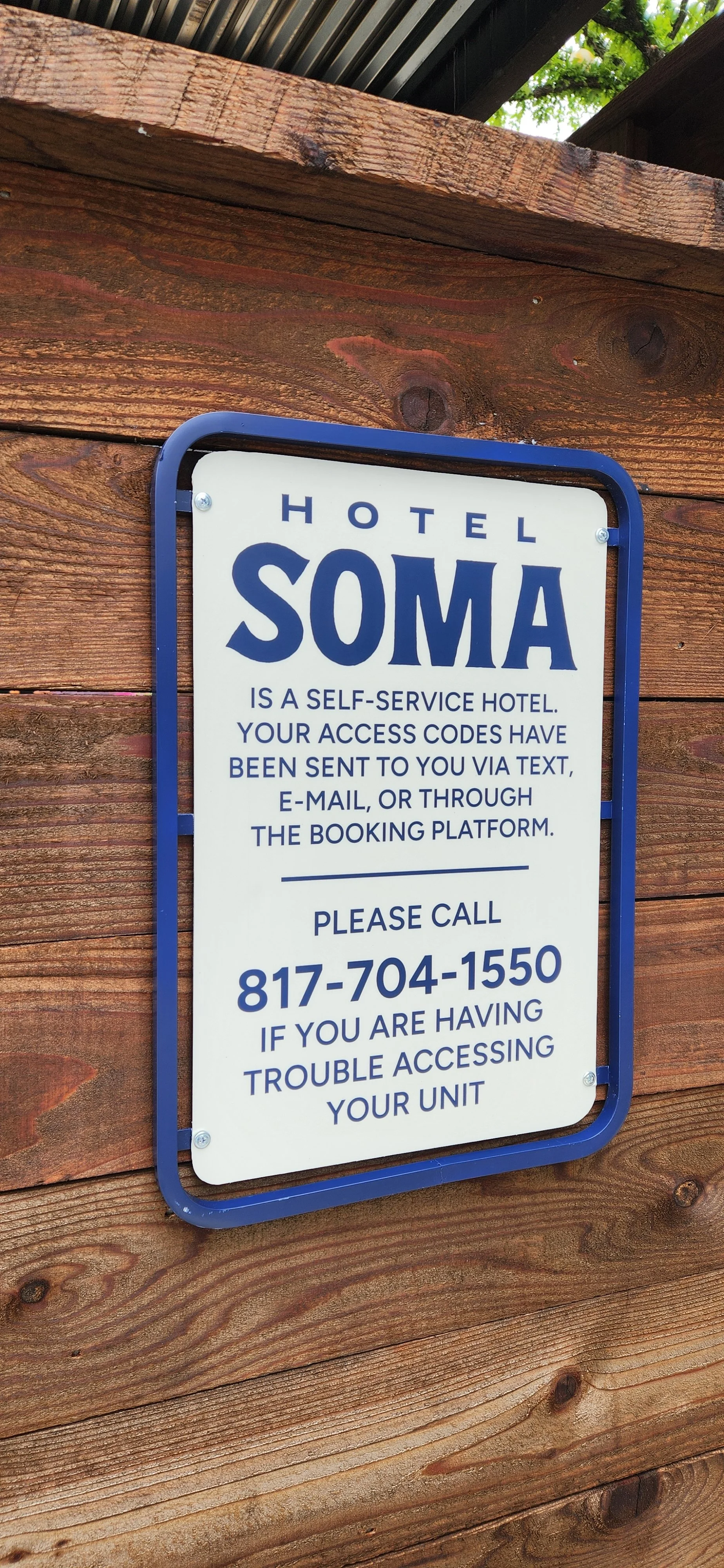 Sign for Hotel Soma explaining it is a self-service hotel with access codes sent via text, email, or booking platform. It provides a phone number for assistance with trouble accessing the unit.