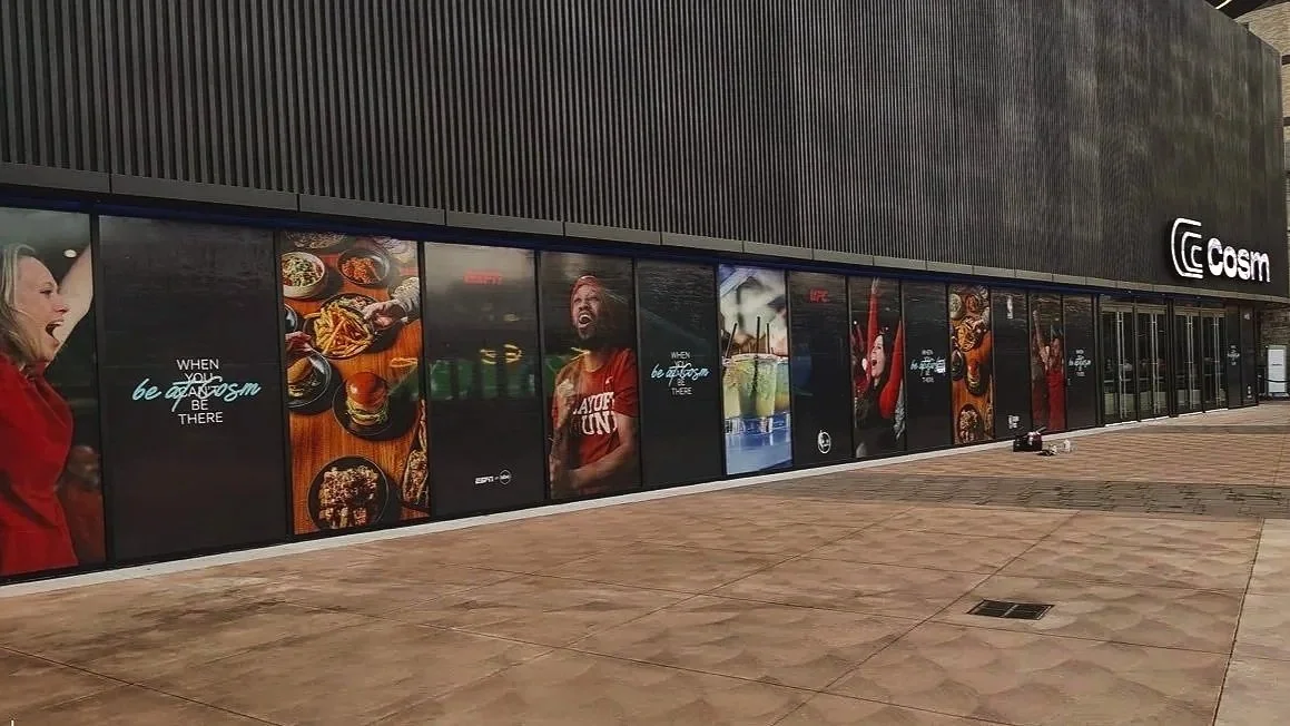 Exterior view of a sports bar or restaurant with large advertisement posters on the windows showcasing food, drinks, and people enjoying themselves, with the logo 'Cosm' on the building.