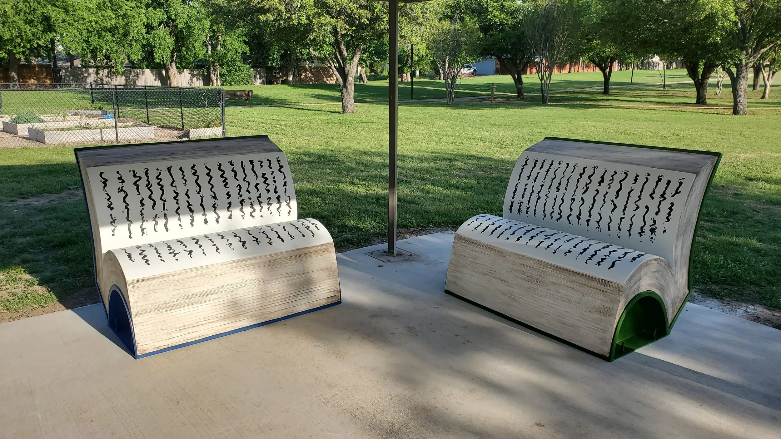 Two modern benches with book design in a park, surrounded by green grass and trees, with a chain link fence and playground area in the background.