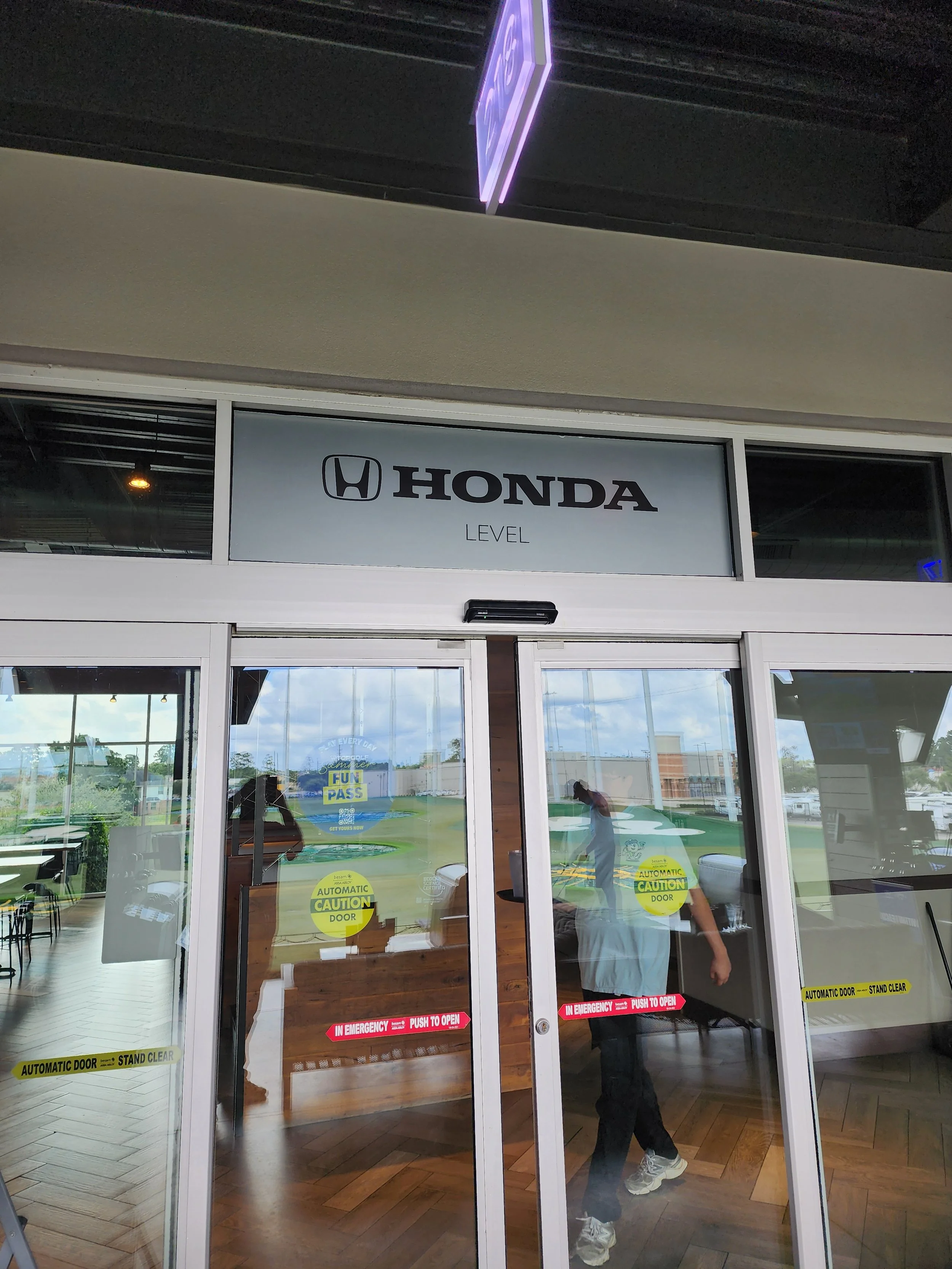 Entrance doors with Honda sign and various caution and automatic door stickers, glass reflecting the inside of a building with a person walking past.