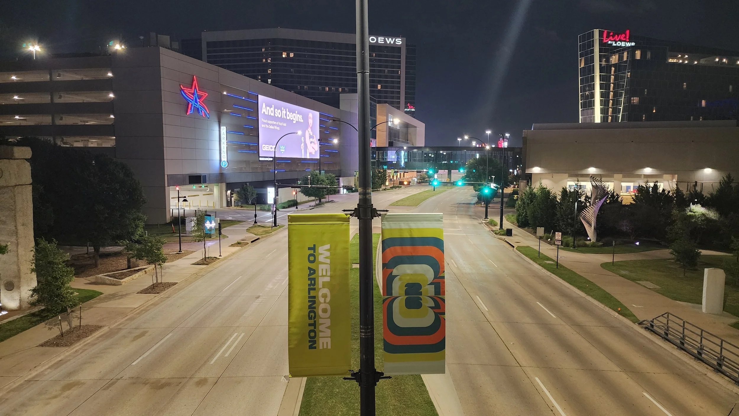 Night view of a city street with empty lanes, surrounded by modern buildings including a mall with electronic billboards, some trees, and streetlights.