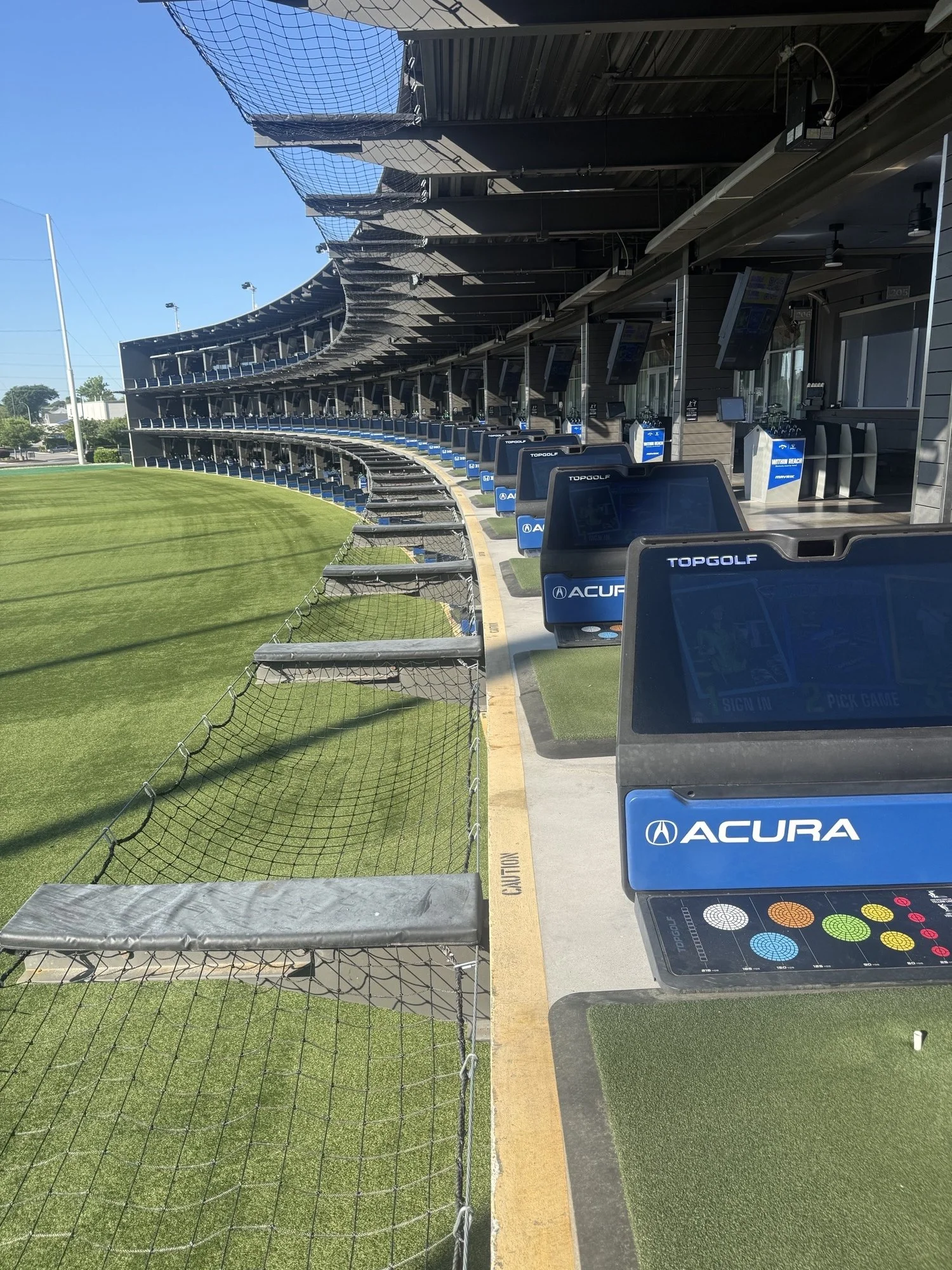 A row of touchscreen kiosks with the Acura logo at a golf driving range, with multiple levels of overhanging nets and a grassy area in front.