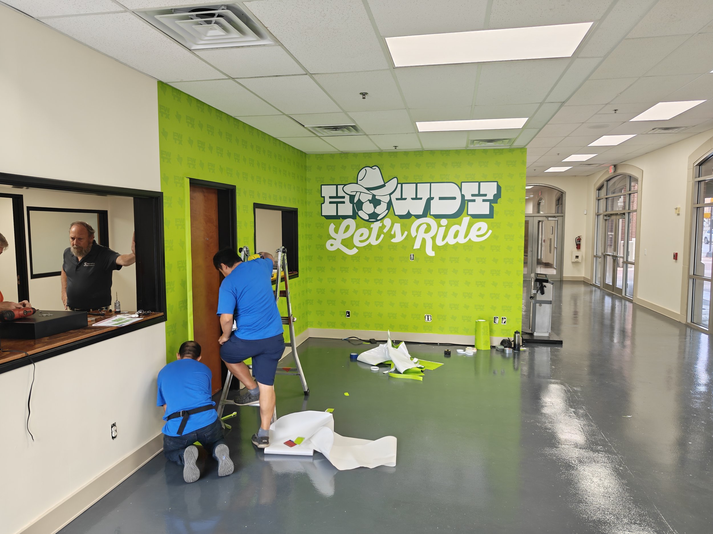 People working on a green wall with the logo "How We Pet Let’s Ride" in an interior space with large windows and ceiling lights.